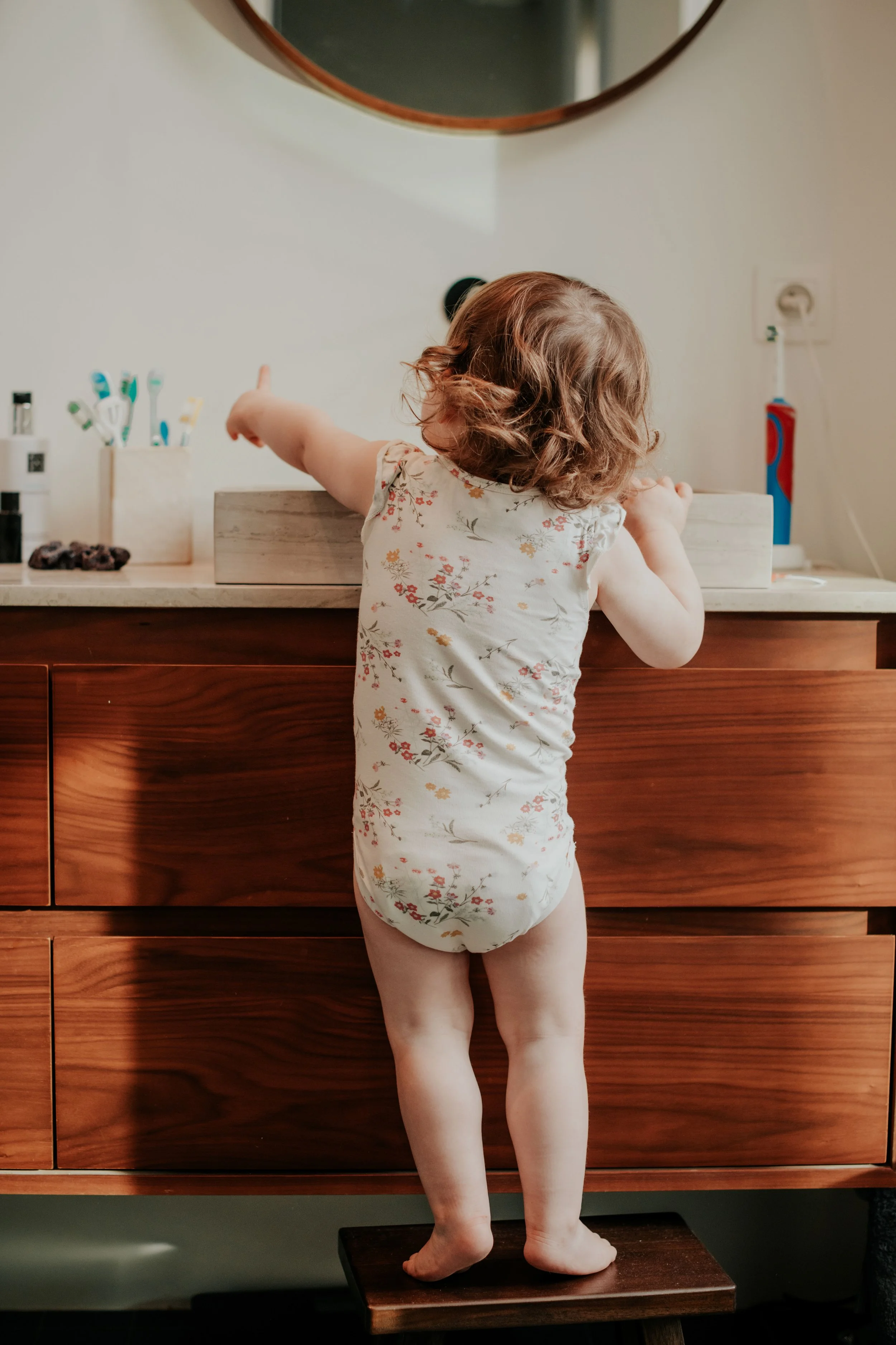A young girl with curly, light brown hair is reaching toward a bathroom mirror on the wall. She is standing on a small wooden step stool and wearing a floral-patterned sleeveless romper. There are toothbrushes and toothpaste on the countertop behind 