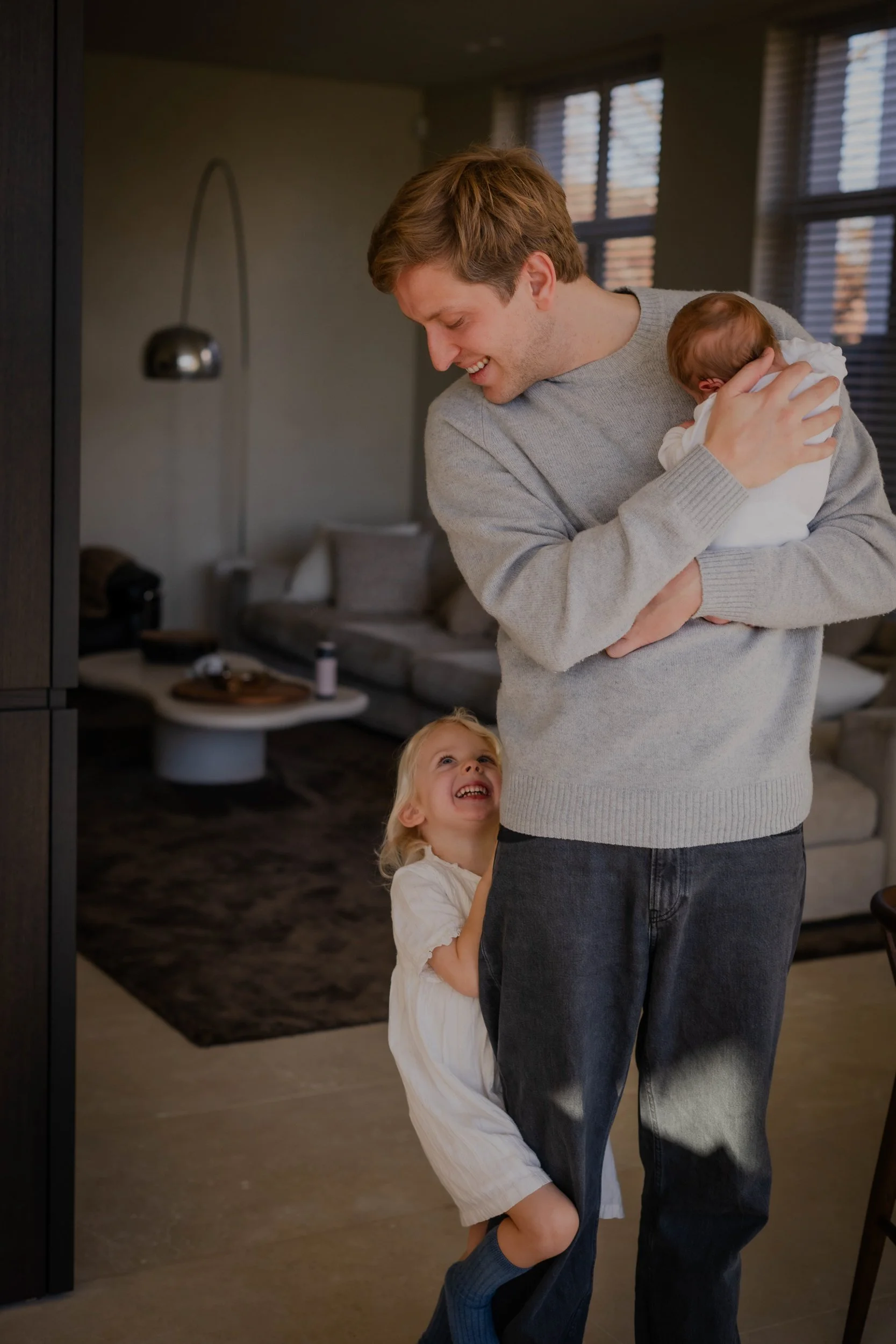A young man in a grey sweater holding a baby girl in white clothing, while a little girl in a white dress and blue socks looks up at them smiling in a living room.