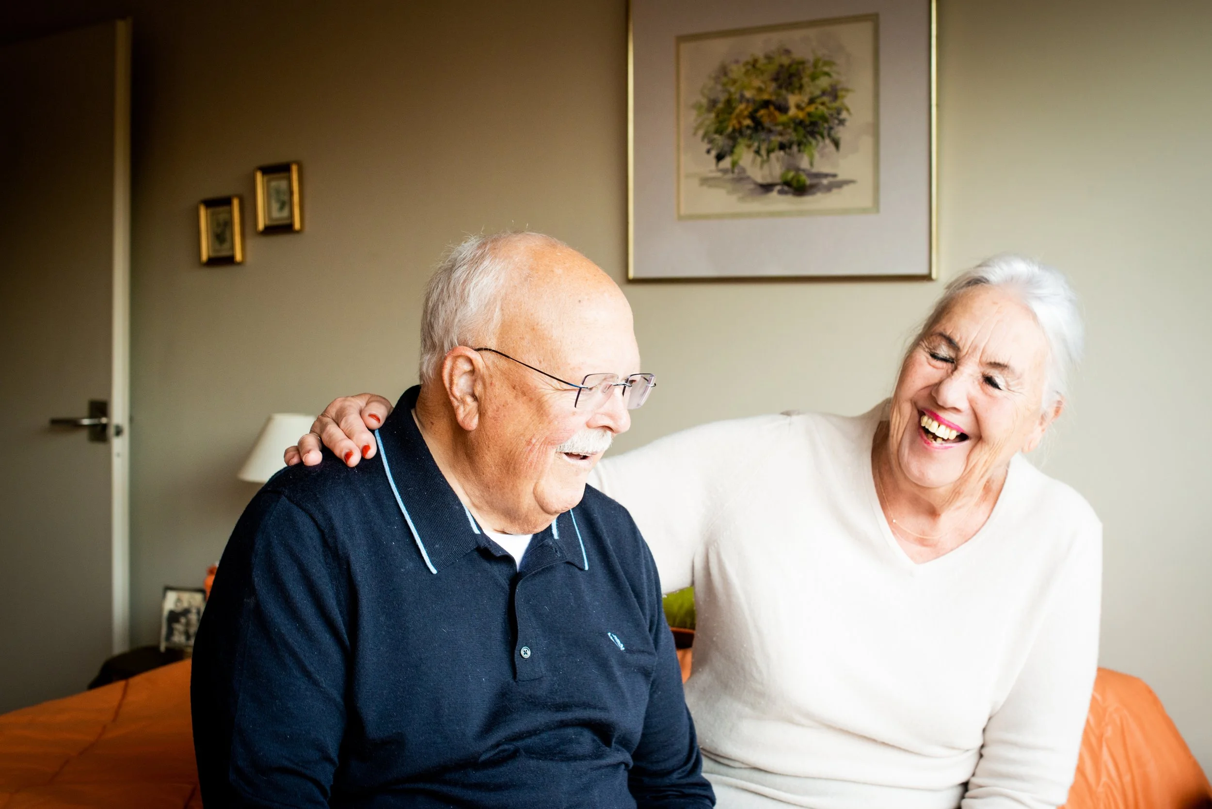 An elderly man and woman are sitting together on a sofa, smiling and laughing, with the woman resting her hand on the man's shoulder. They appear to be enjoying each other's company in a cozy living room.