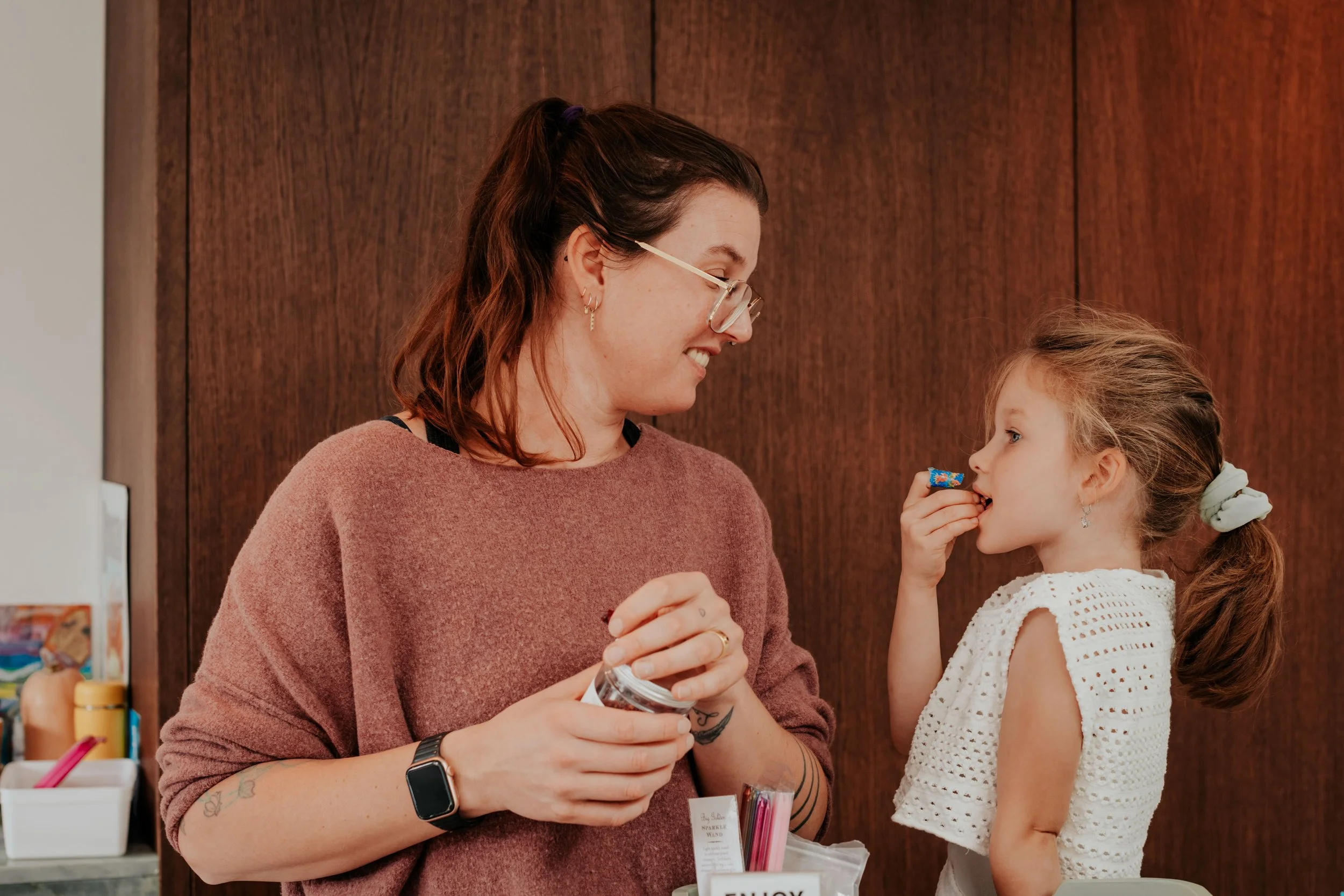 A woman and a young girl sharing a moment, with the woman holding a jar of candy and the girl reaching in to get some, in an indoor setting.