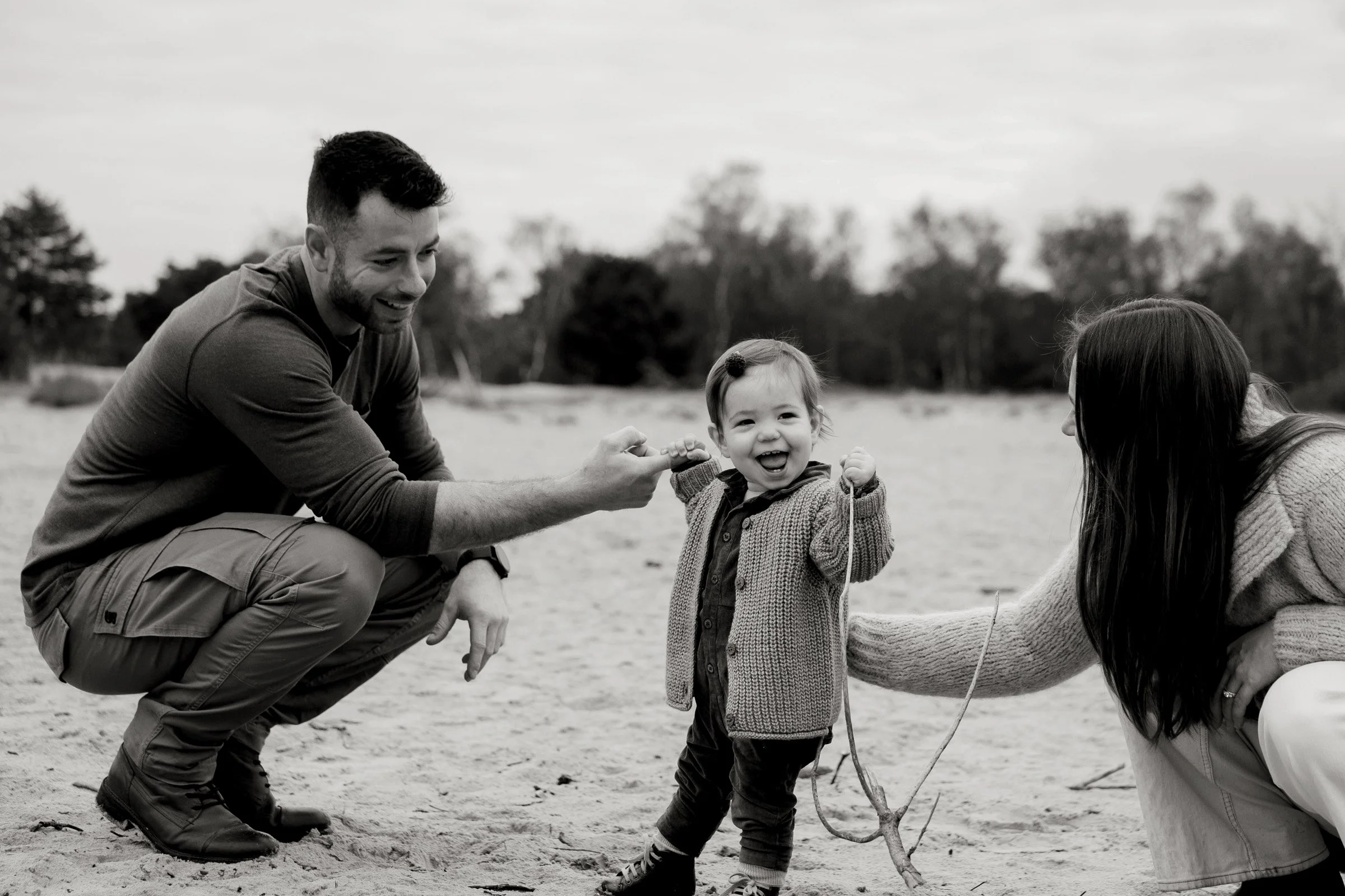 A man and a woman helping a young girl hold a stick in a sandy outdoor setting, with trees in the background.