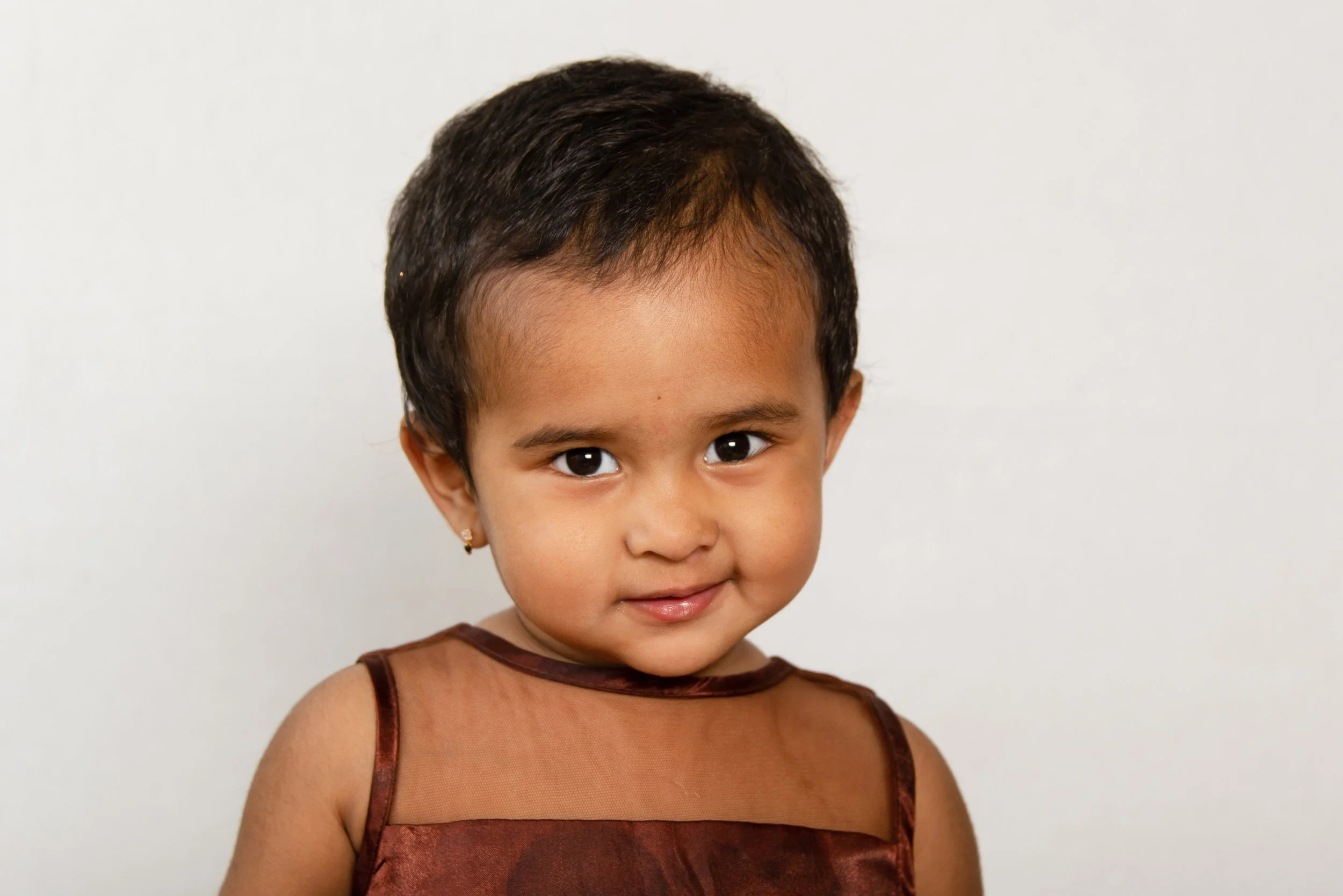 Close-up of a young girl with dark hair and large brown eyes wearing a brown sleeveless top against a plain white background.