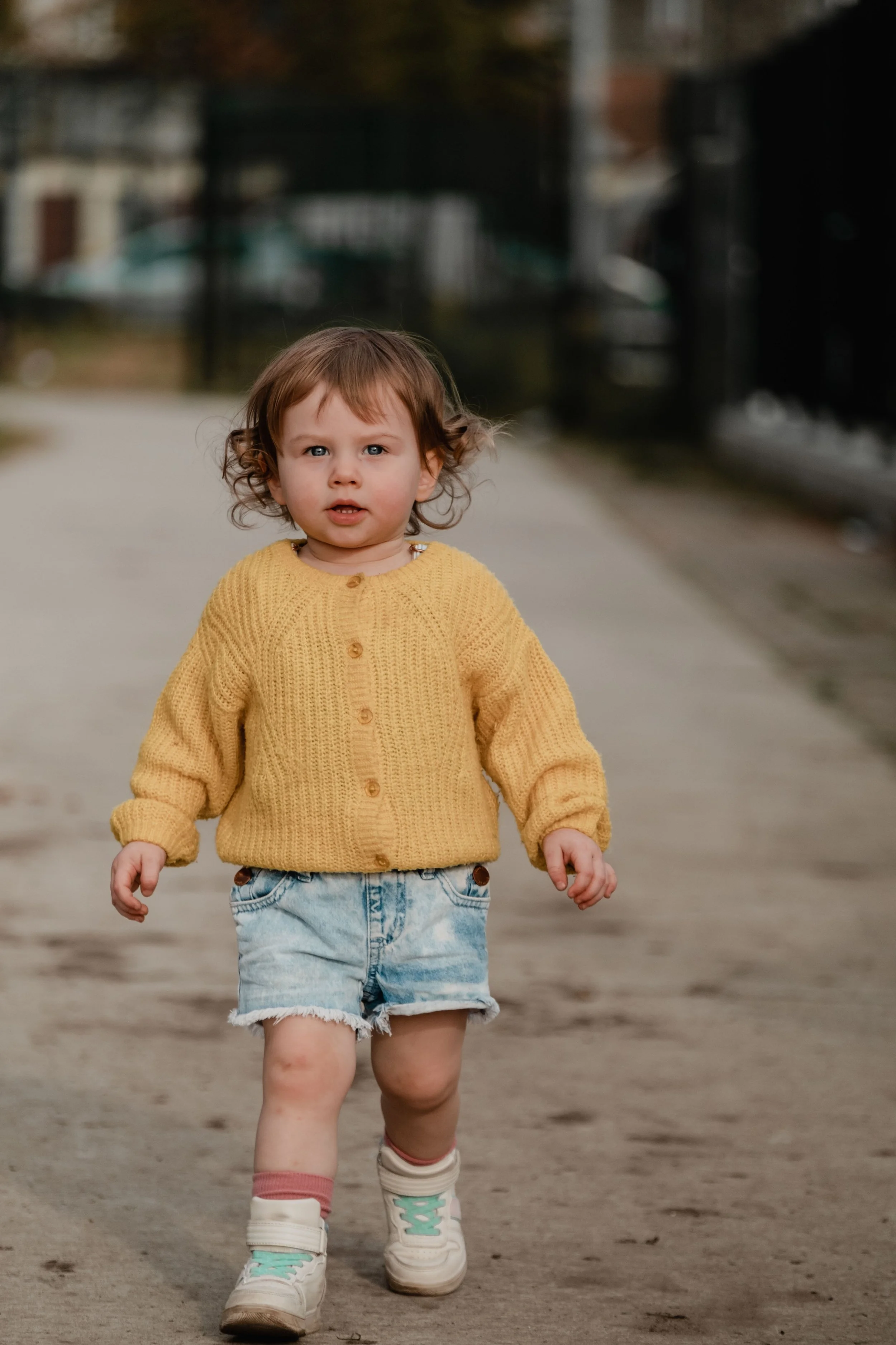 A young child with curly brown hair, wearing a yellow knitted sweater, denim shorts, pink socks, and white sneakers, walking on a dirt path outdoors.