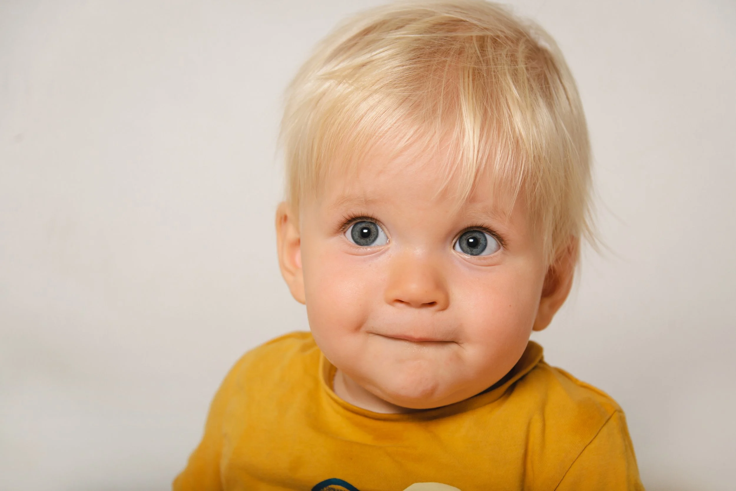 Close-up of a young child with blonde hair and blue eyes, wearing a yellow shirt and looking directly at the camera.