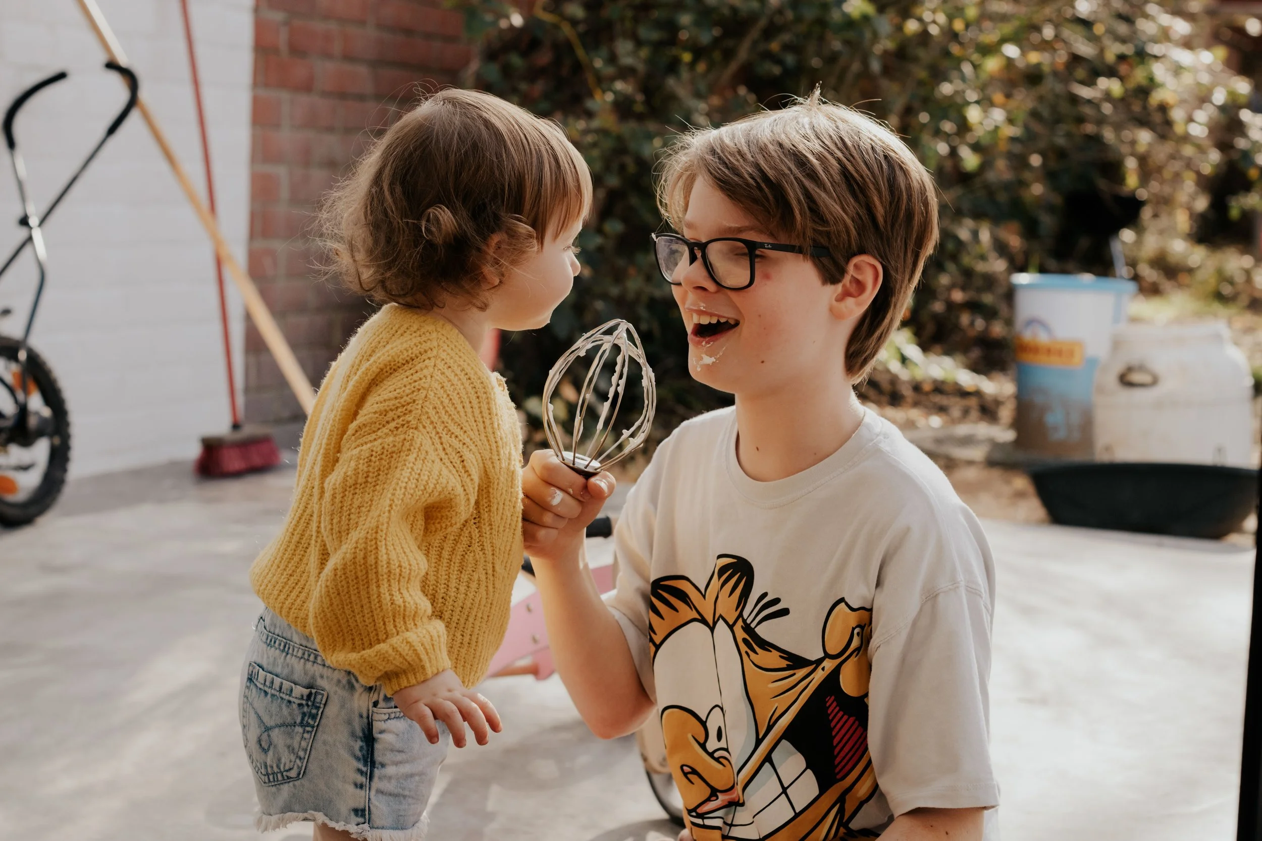A young boy with glasses and a white shirt is smiling and holding a wire whisk, while a little girl in a yellow sweater and denim shorts leans in close for a playful moment outside.