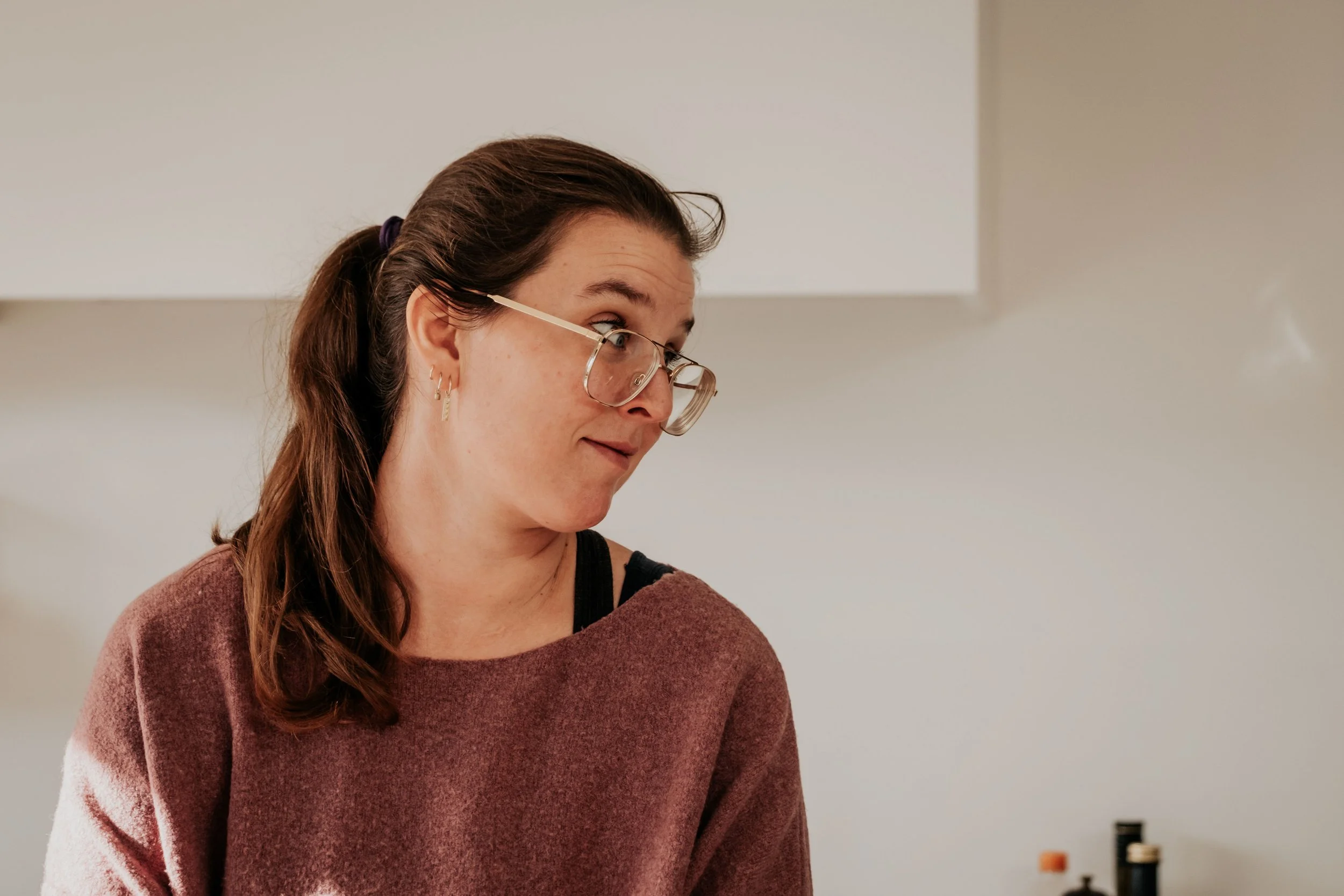 A woman with brown hair tied back wearing glasses and earrings, looking down, wearing a maroon sweater in a room with a white wall.
