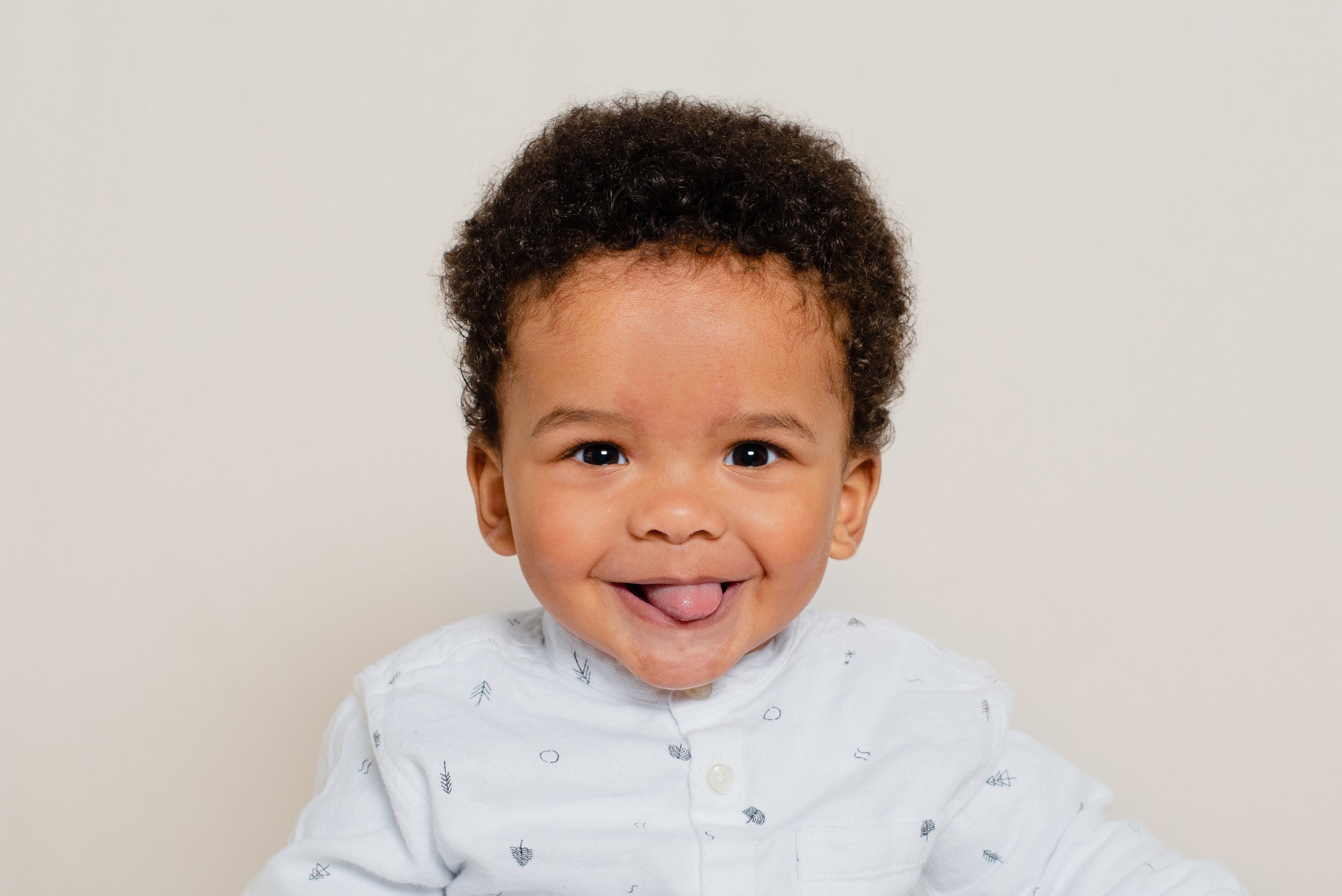Close-up of a smiling toddler with curly hair, sticking out tongue, wearing a white shirt with small black patterns, against a plain light background.