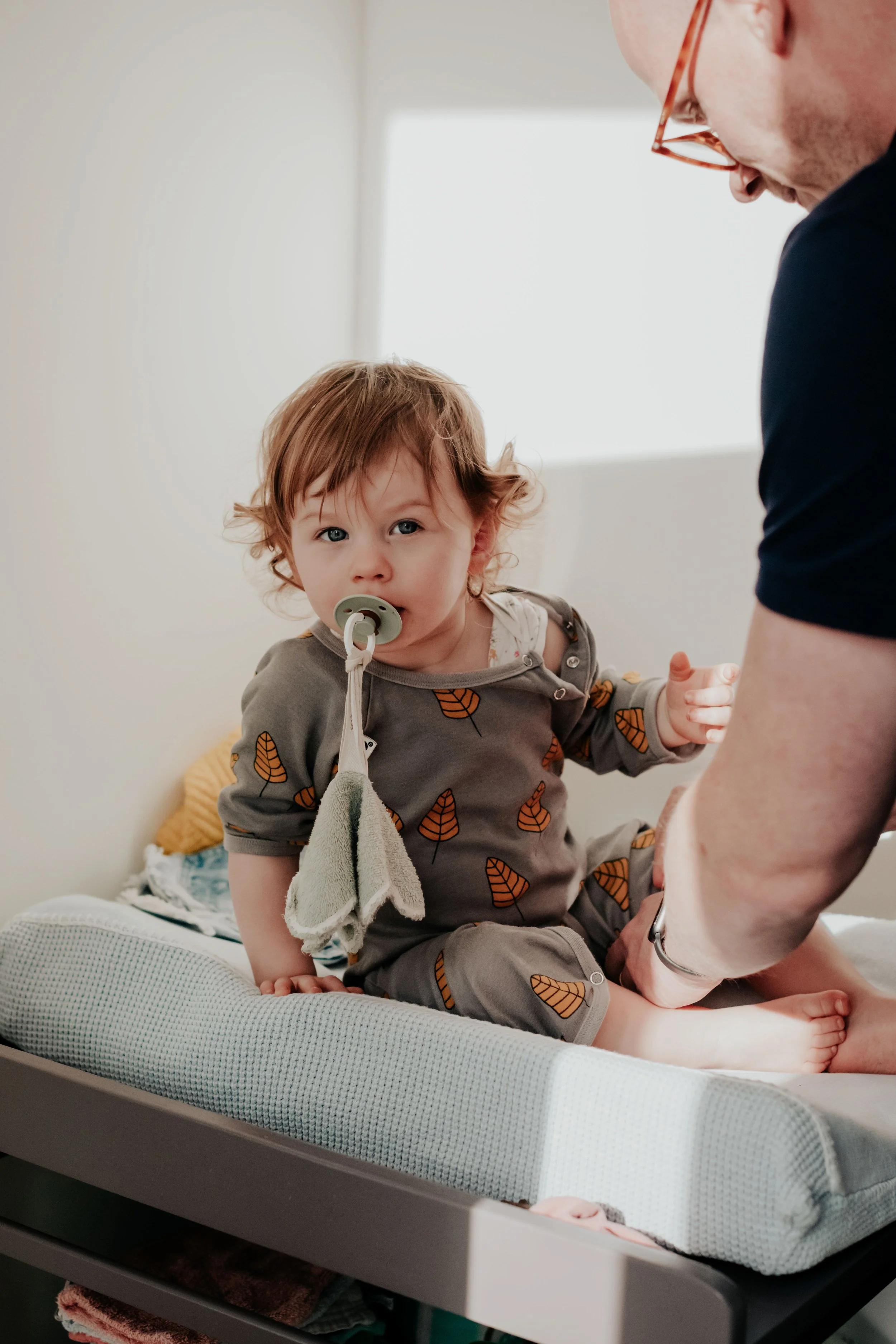 A young child with red hair and a pacifier in their mouth, sitting on a changing table with a woman beside them, during a diaper change in a bright room.