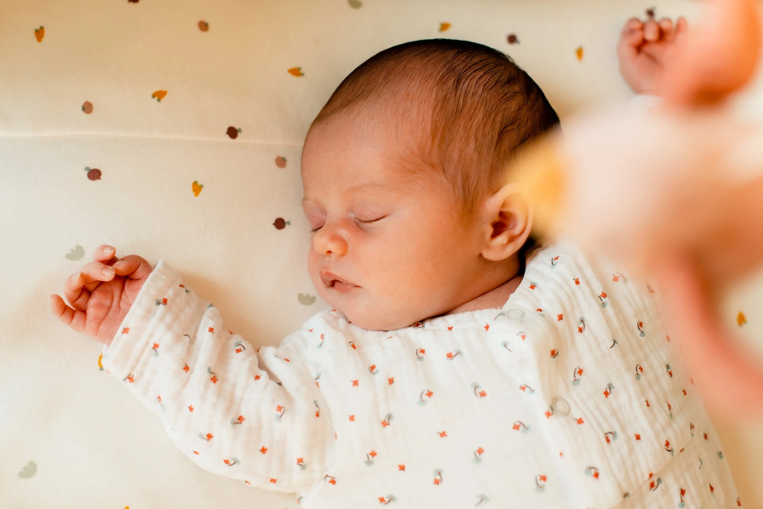 A sleeping newborn baby baby lying on a white surface with small colored fruit illustrations, dressed in a white outfit with tiny orange and black pattern, holding one hand up.