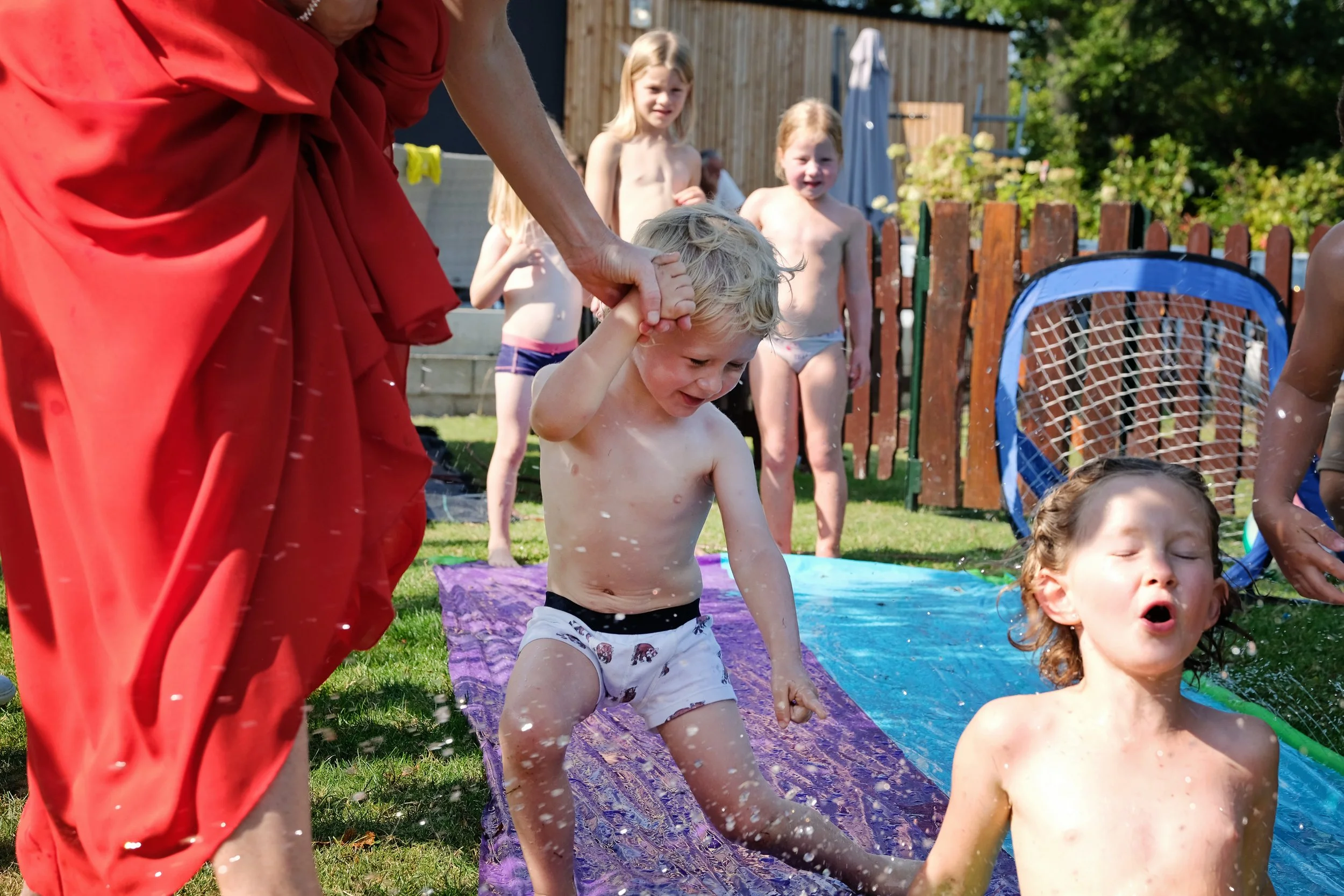 Children and an adult playing water games on a sunny outdoor day, with grass, a wooden fence, and trees in the background.