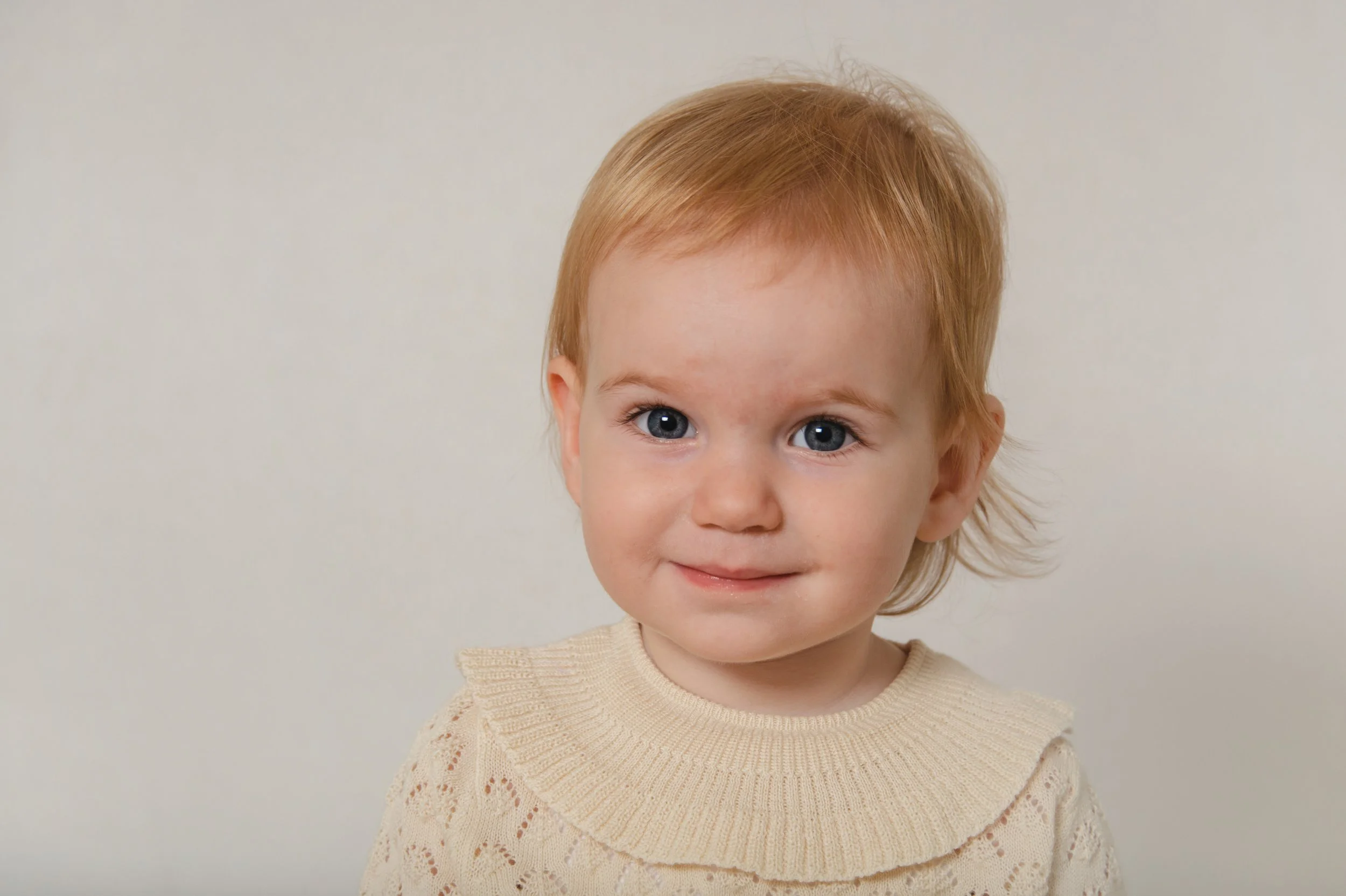 A young child with red hair and bright blue eyes smiling at the camera, wearing a cream-colored knit sweater, against a plain off-white background.