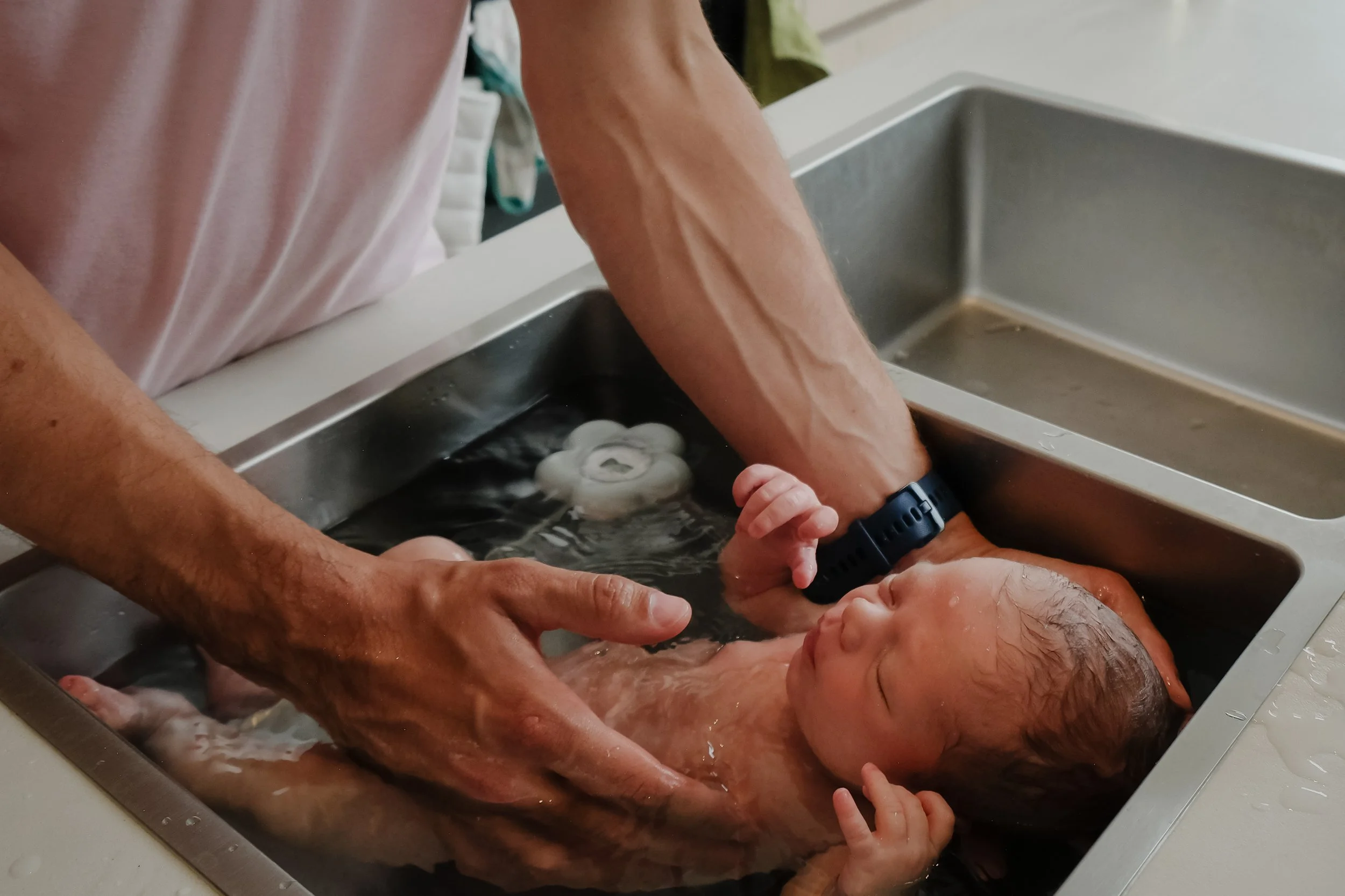 A person with a watch on their wrist is giving a bath to a newborn baby in a sink, with multiple hands helping during the process.