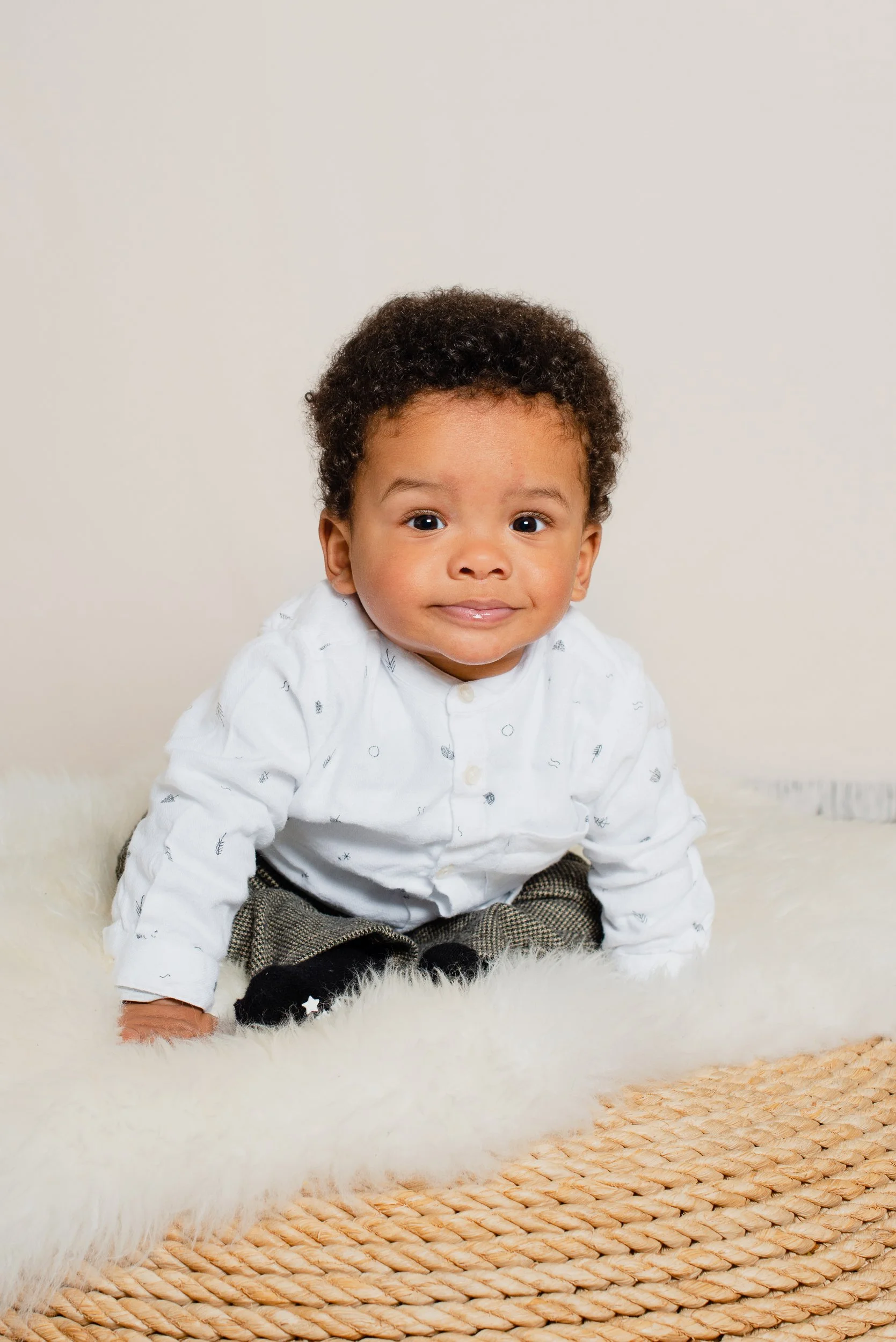 Young child with curly hair and light brown skin, sitting on a fuzzy white rug in front of a plain light-colored wall, wearing a white shirt with small patterns, plaid pants, and black socks with stars, smiling at the camera.