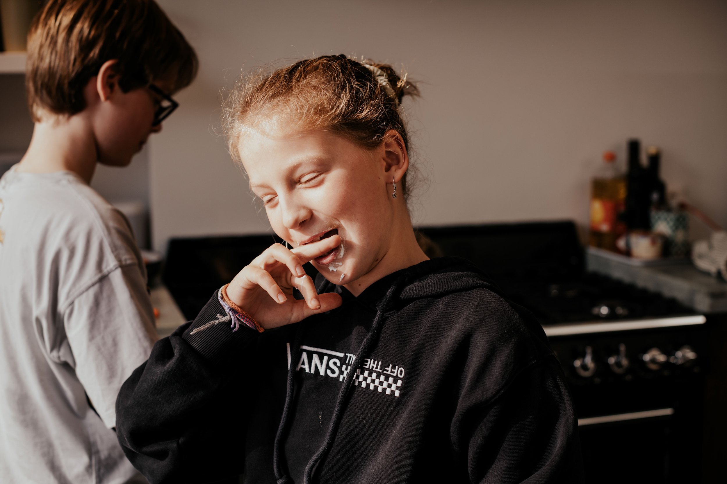 A young woman with red hair in a ponytail smiling with her eyes closed as she touches her face, wearing earrings and a black hoodie, while a person with short brown hair and glasses stands nearby in a room with a keyboard and some bottles in the back