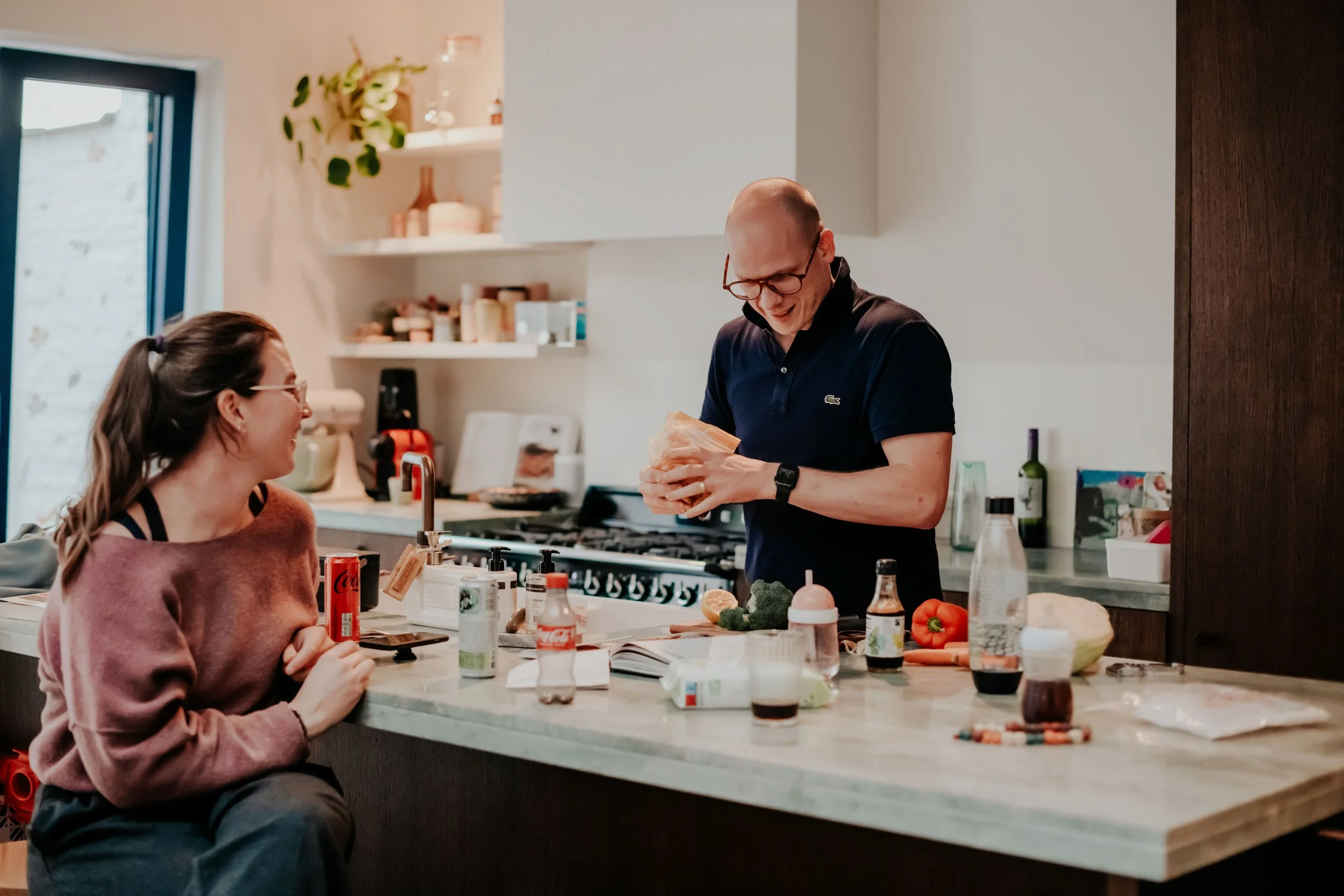 A woman and a man are in a kitchen, smiling and talking. The woman is sitting at a white kitchen island, wearing glasses and a pink sweater. The man, wearing glasses and a dark blue shirt, is holding a package and standing in front of the island. The