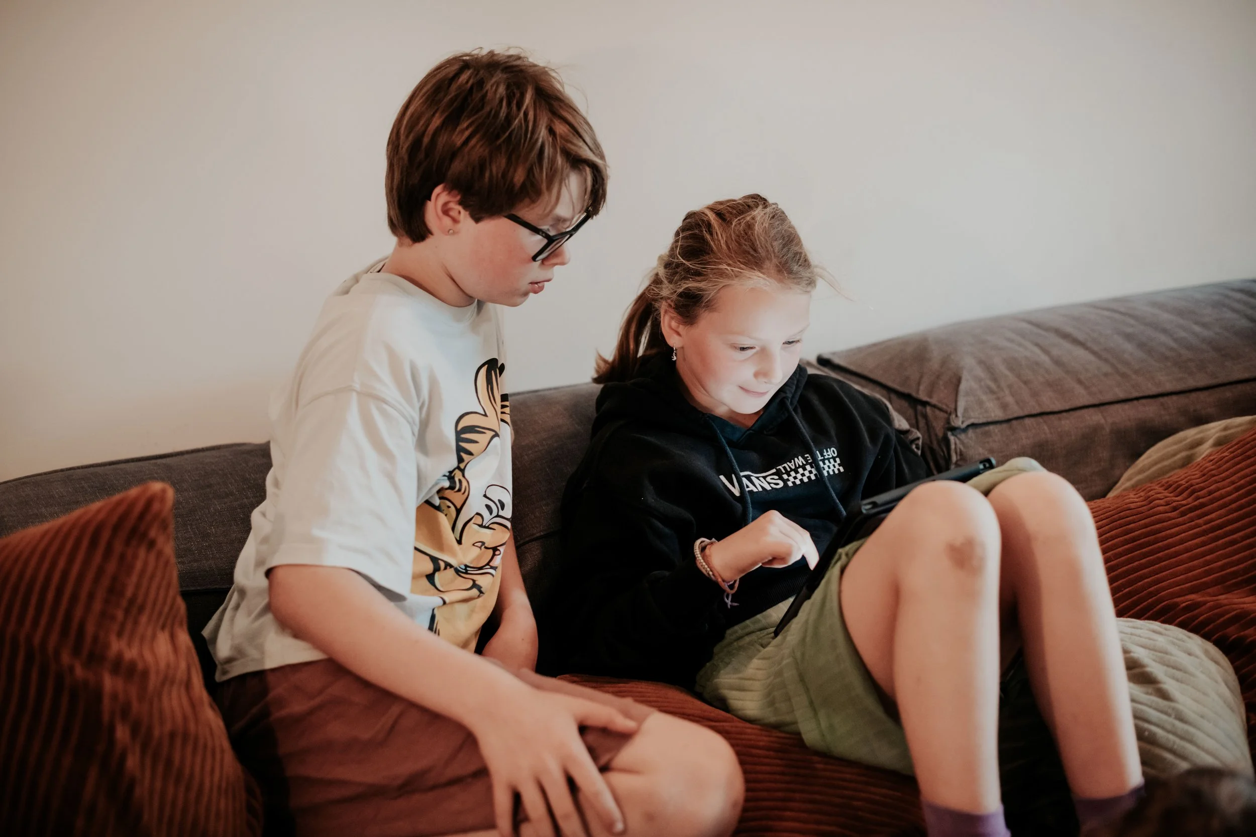 Two children, a boy and a girl, sitting on a couch, looking at a phone together.