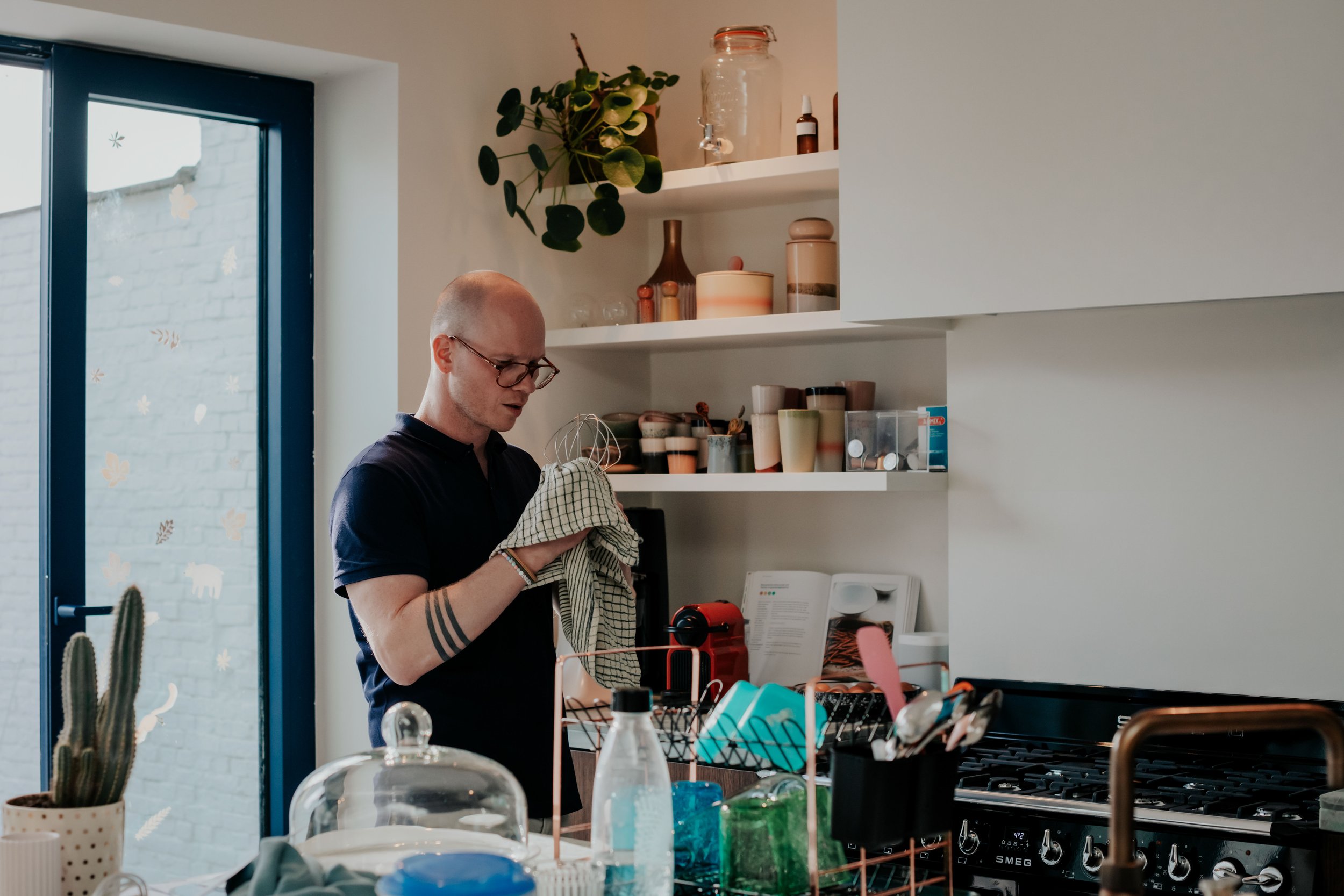 A man with glasses and tattoos holding a dish towel in a kitchen filled with various objects including plants, bottles, and containers, near a window with decorative stickers.