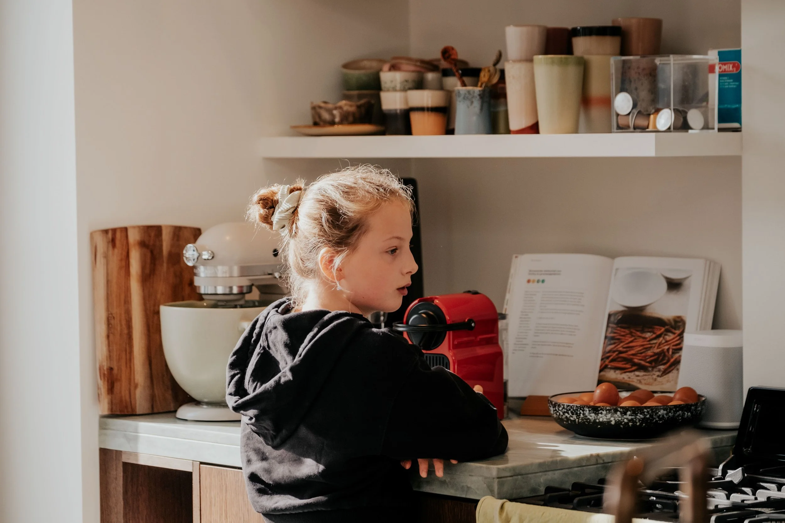 A girl with blonde hair tied in a bun, wearing a black hoodie, sitting at a kitchen counter reading a magazine with a bowl of eggs nearby.