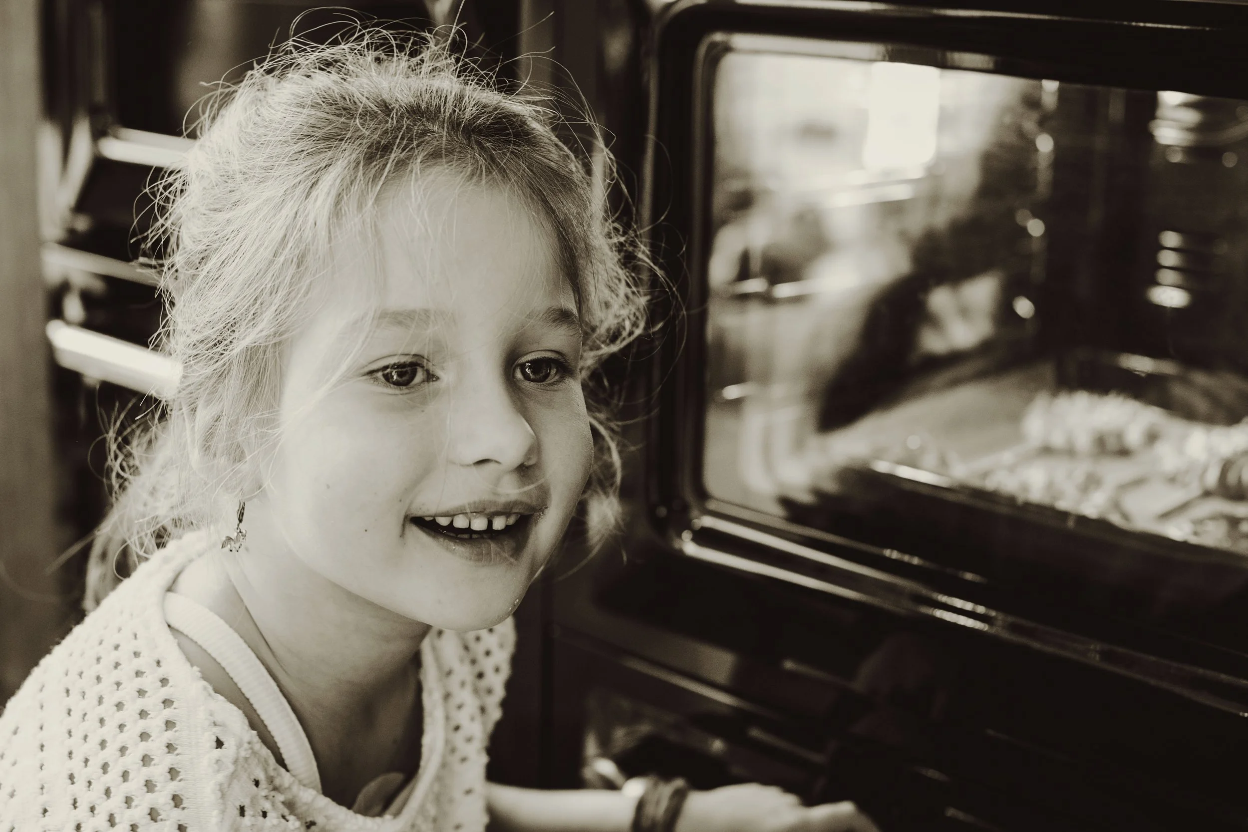 A young girl with curly hair smiling as she looks into an oven with baked food inside.
