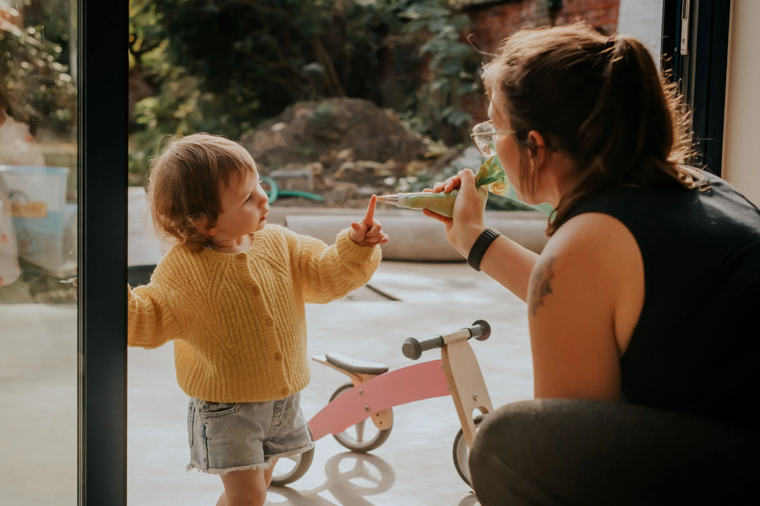 A young child with light brown hair, wearing a yellow sweater and denim shorts, points at an adult woman's nose while standing outside near a large window. The woman, with brown hair tied back, is holding a green bag and is leaning lower towards the 