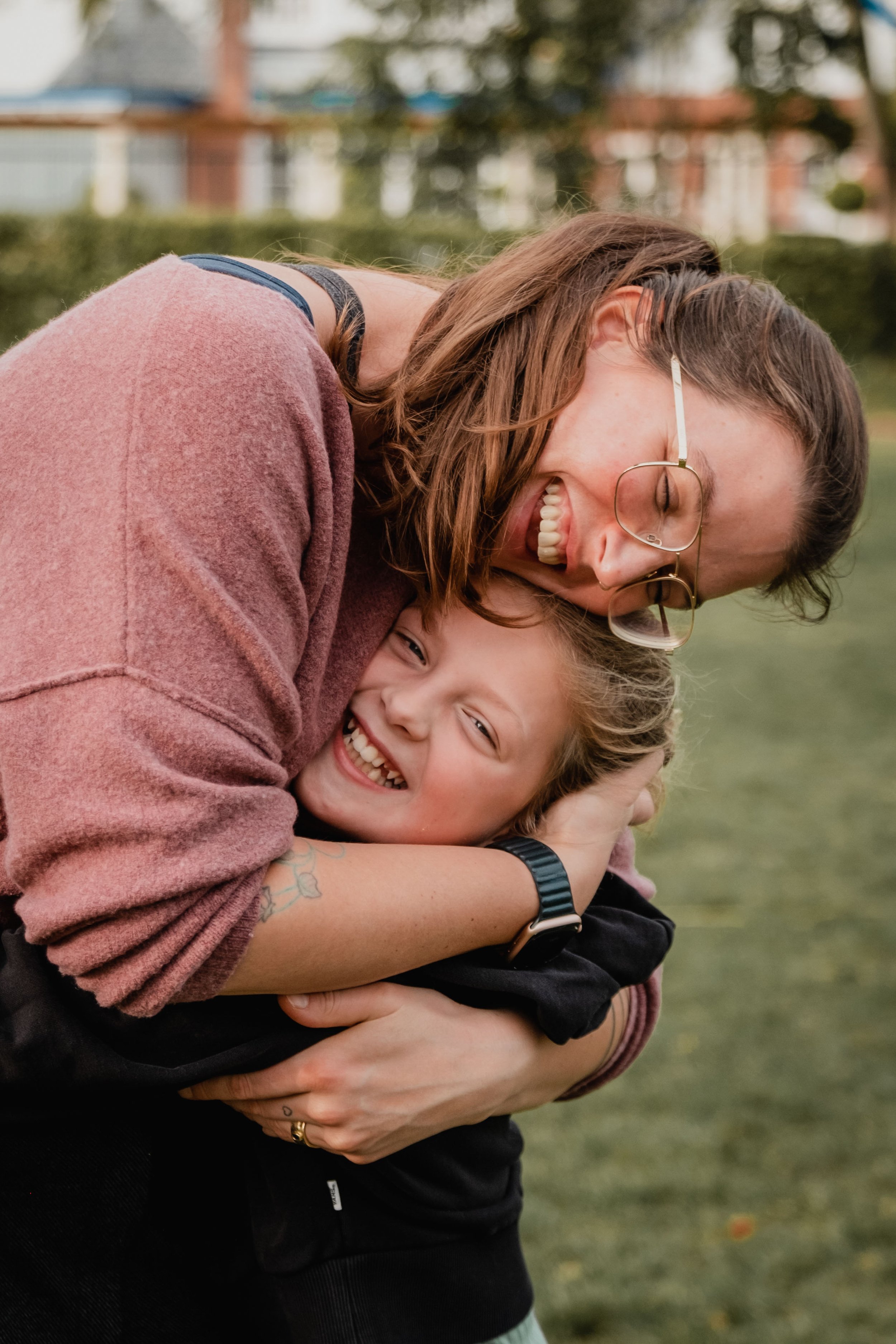 A woman with glasses and long brown hair hugging a smiling child with blonde hair in an outdoor setting with trees and houses in the background.