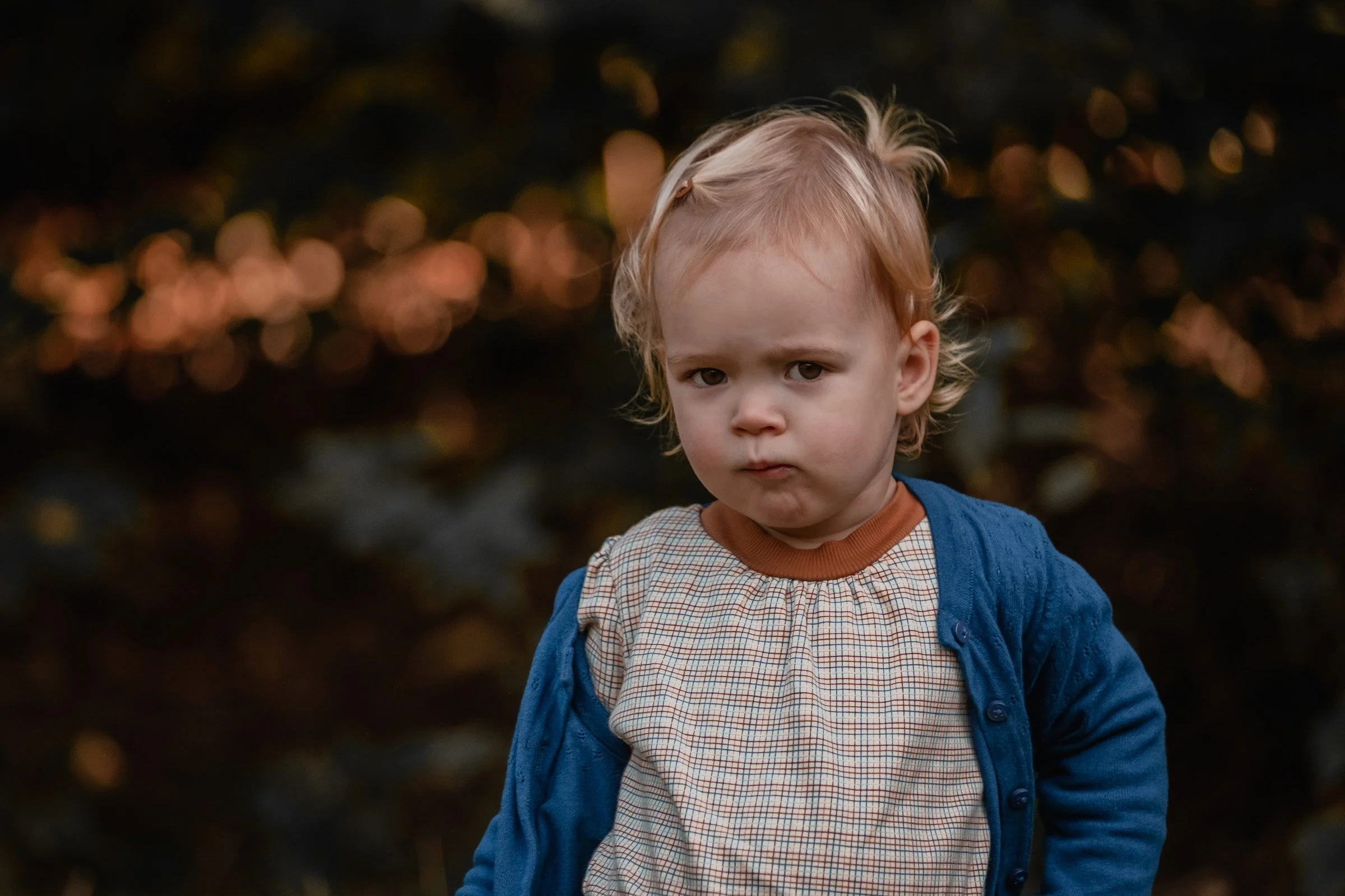 A young child with blonde hair and a serious expression, wearing a checkered shirt and a blue jacket, standing outdoors at dusk with blurred lights in the background.