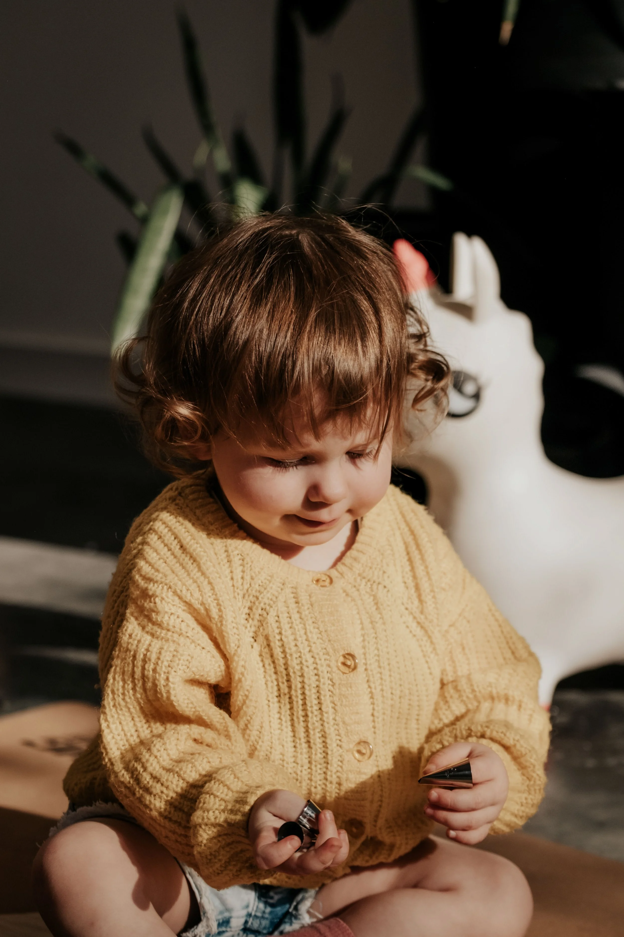 A young child with curly brown hair, wearing a mustard-colored cardigan and shorts, sits cross-legged indoors, playing with toy cars. In the background, there is a large white sculptural sculpture and a tall green plant with multiple leaves.