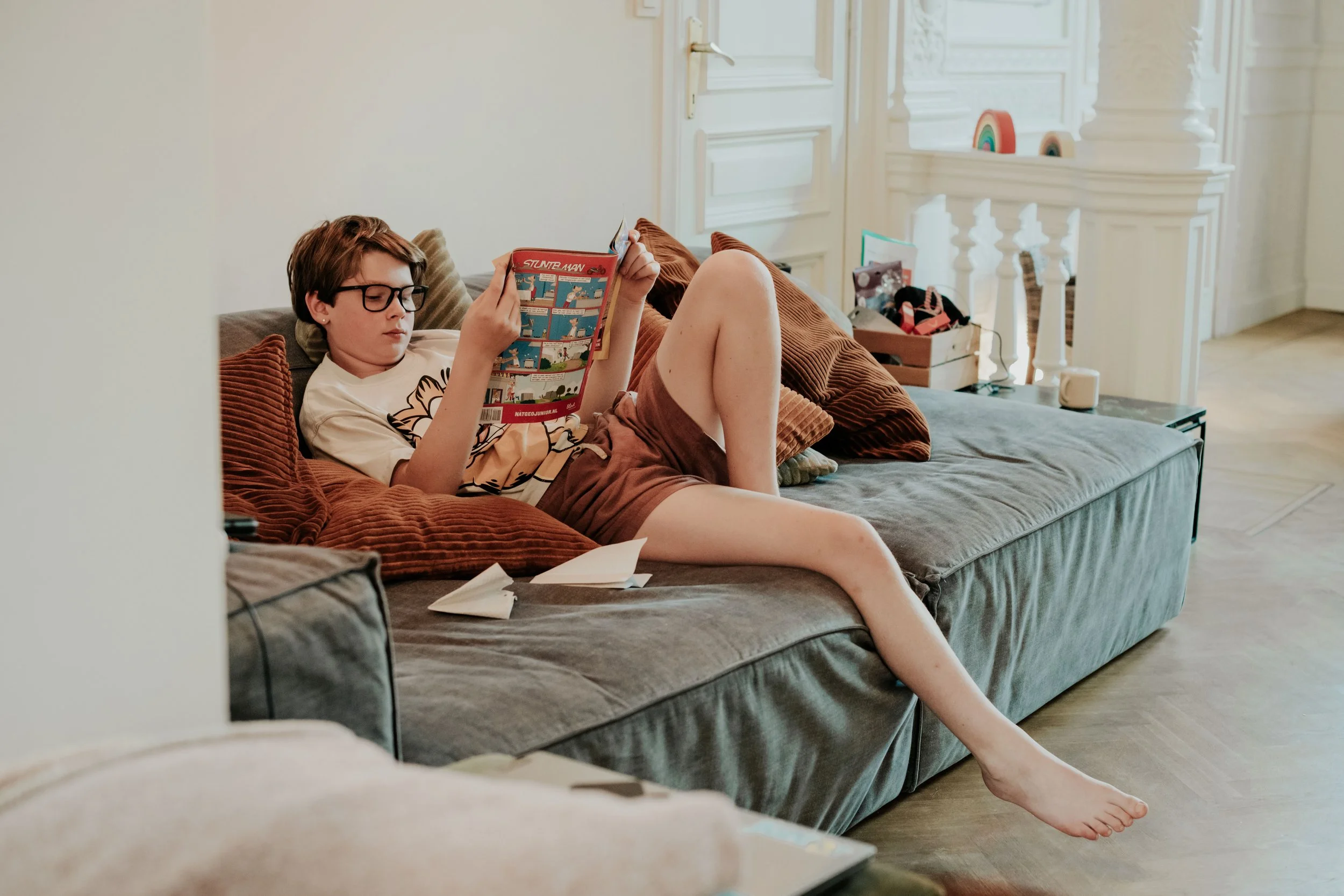A young person with short brown hair and glasses is lying on a brown couch, reading a comic book. They are wearing a light-colored T-shirt with a graphic design and brown shorts. The room has white walls, decorative molding, and a wooden floor. There