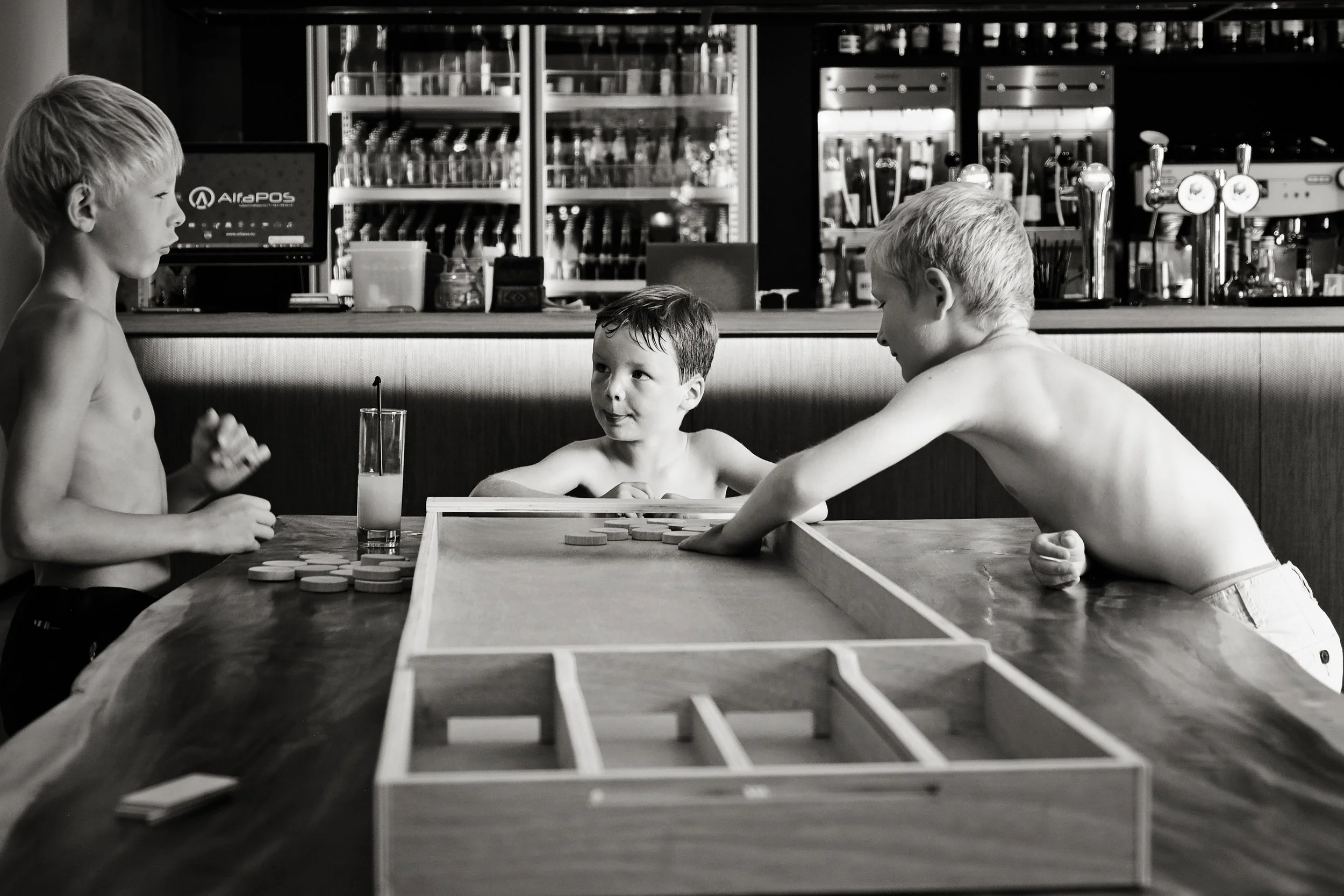 Three young boys playing a tabletop game with wooden tokens in a café or bar setting. The background features shelves with bottles and bar equipment. The image is in black and white.