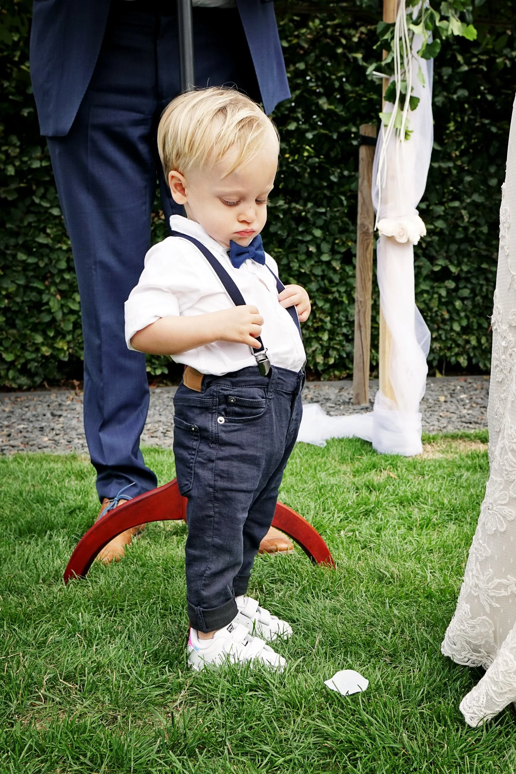 Young boy dressed in a white shirt, bow tie, and black pants is standing outdoors on green grass, holding suspenders. Behind him, there is a man in a dark suit with his face not visible, and decorative white fabric with flowers and greenery. The scen
