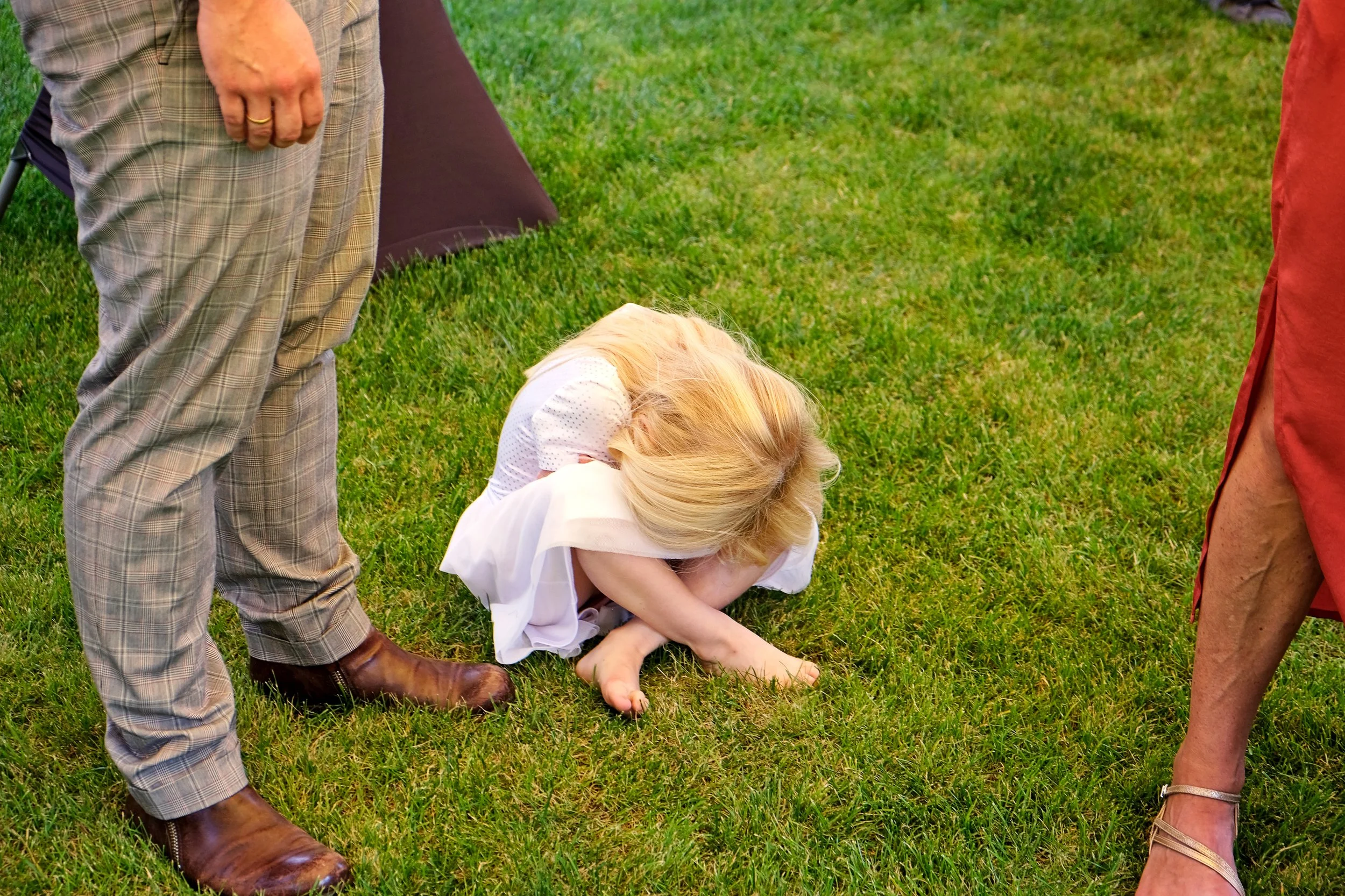 Child with long blonde hair lying on grass, surrounded by adults during a social gathering.