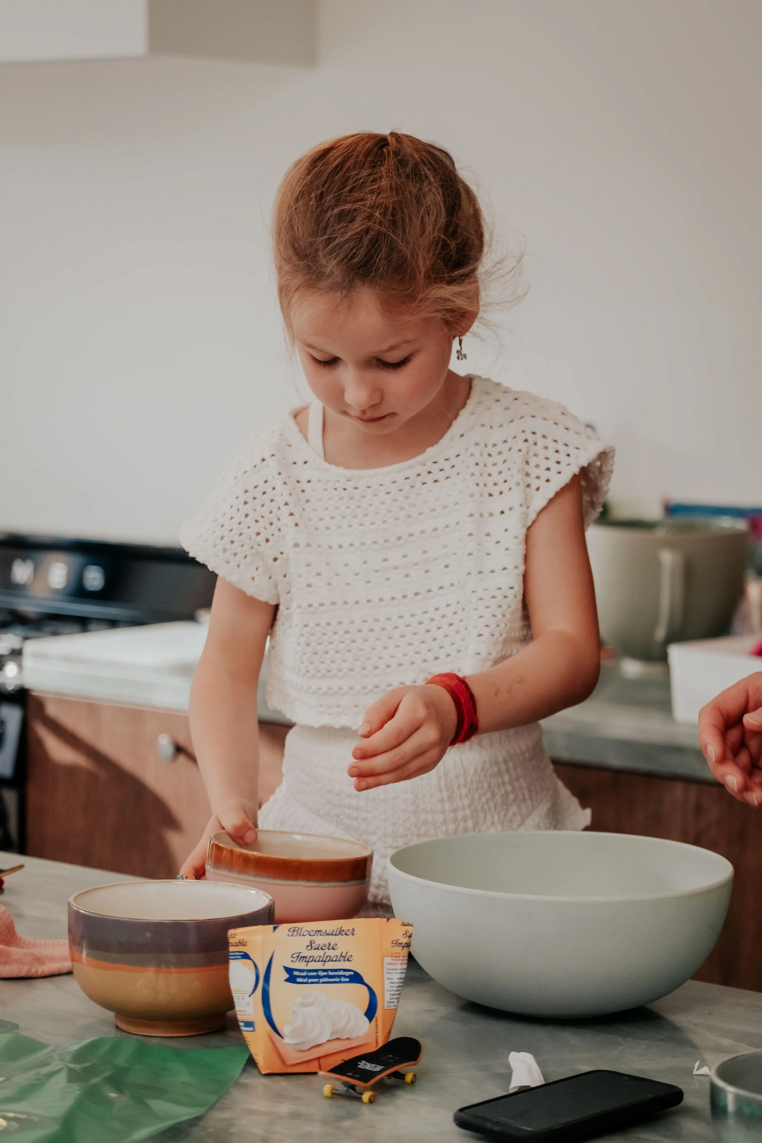 A young girl with red hair and earrings is focused on an activity, standing at a kitchen counter with bowls, a bag of whipped topping, and a skateboard in front of her.