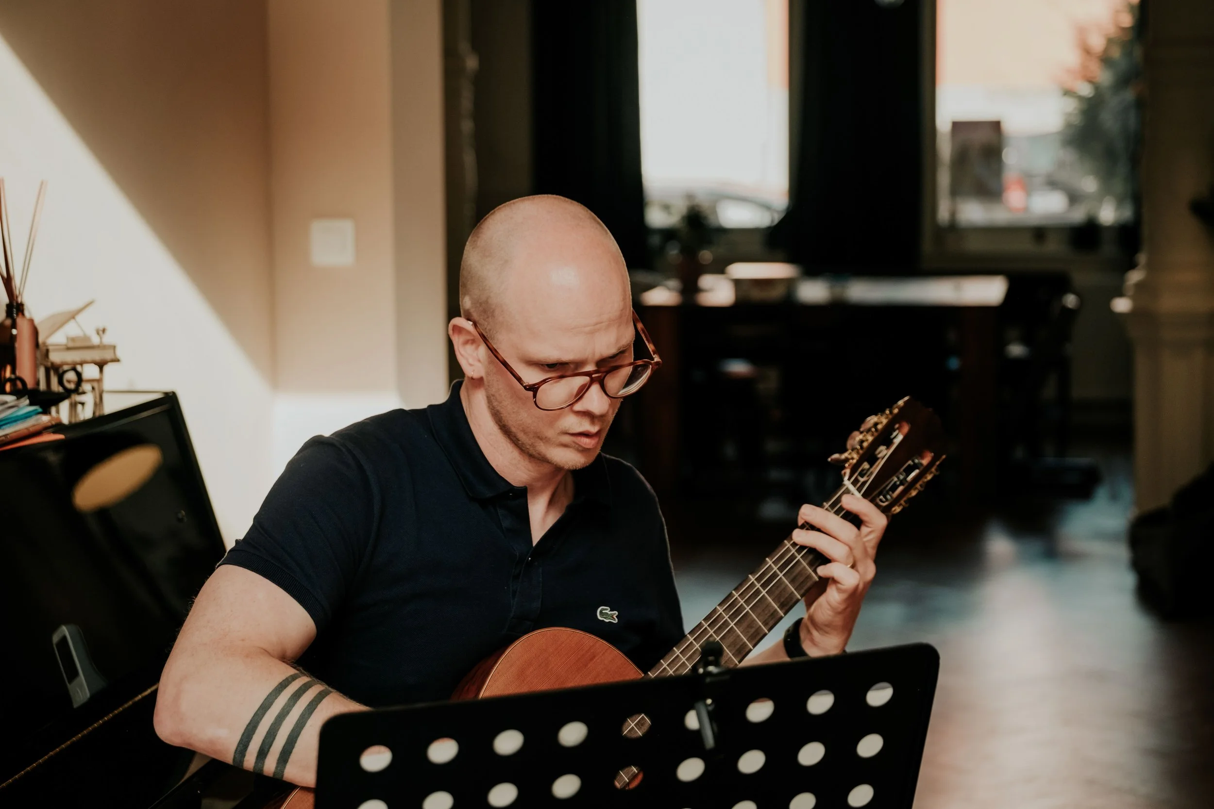 A man with glasses playing an acoustic guitar indoors, seated at a music stand, with a music studio background, sunlight coming through windows.