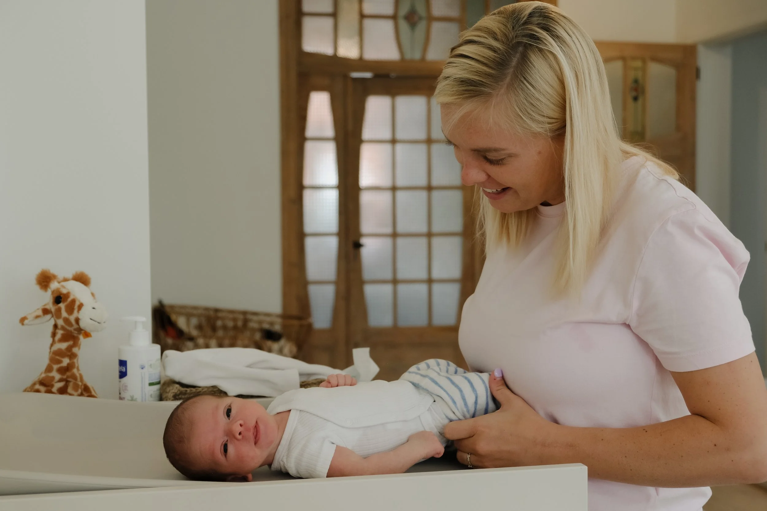 A smiling blonde woman in a pink shirt holding a newborn baby dressed in white and blue stripes. The baby is lying on a changing table with a stuffed giraffe toy nearby and baby supplies on the side. The room has a wooden door with glass panels in th