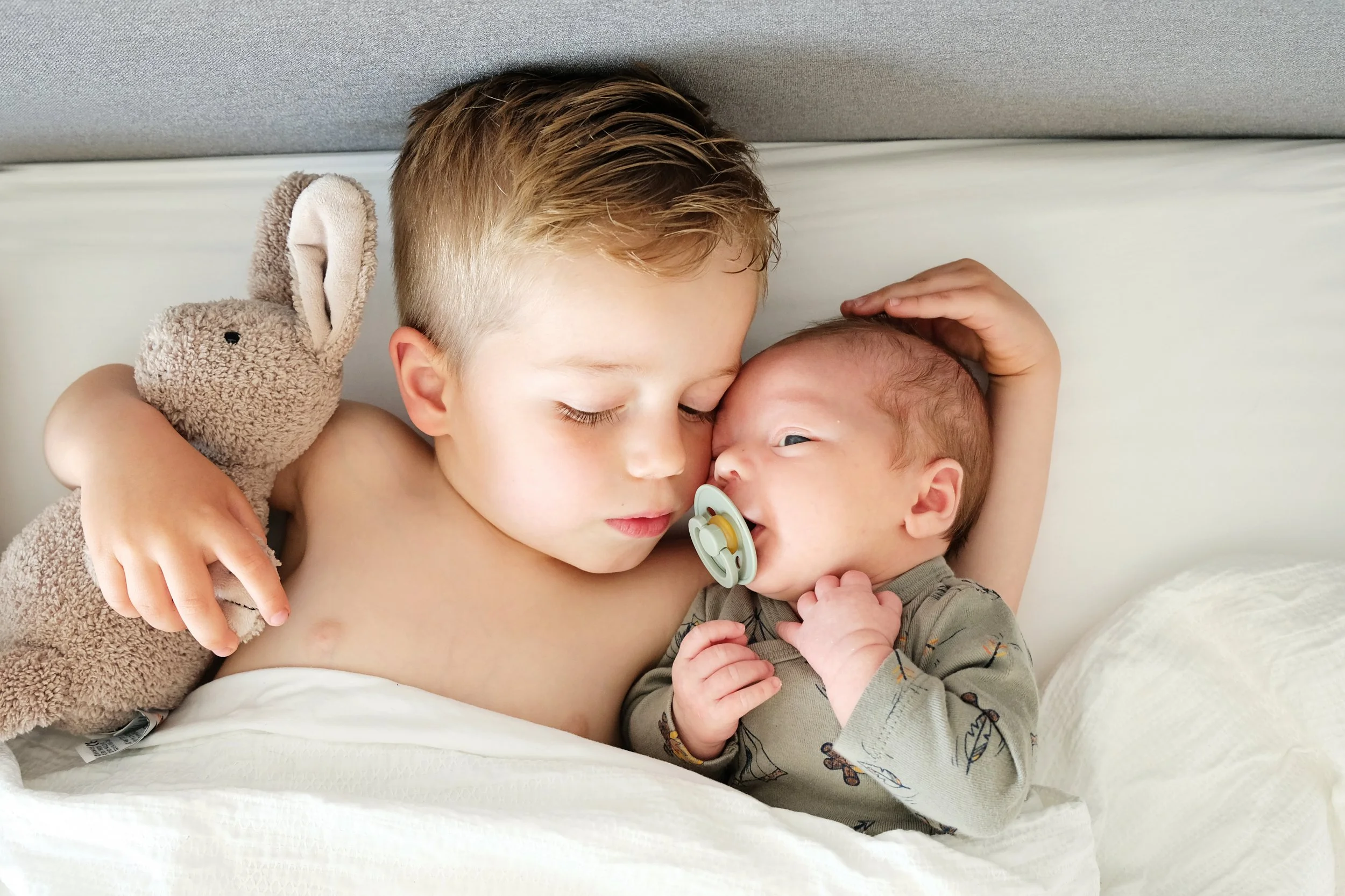 A young boy with light brown hair holding a newborn with a pacifier in their mouth, lying together on a bed. The boy has a plush bunny toy and gentle hand on the baby's head.