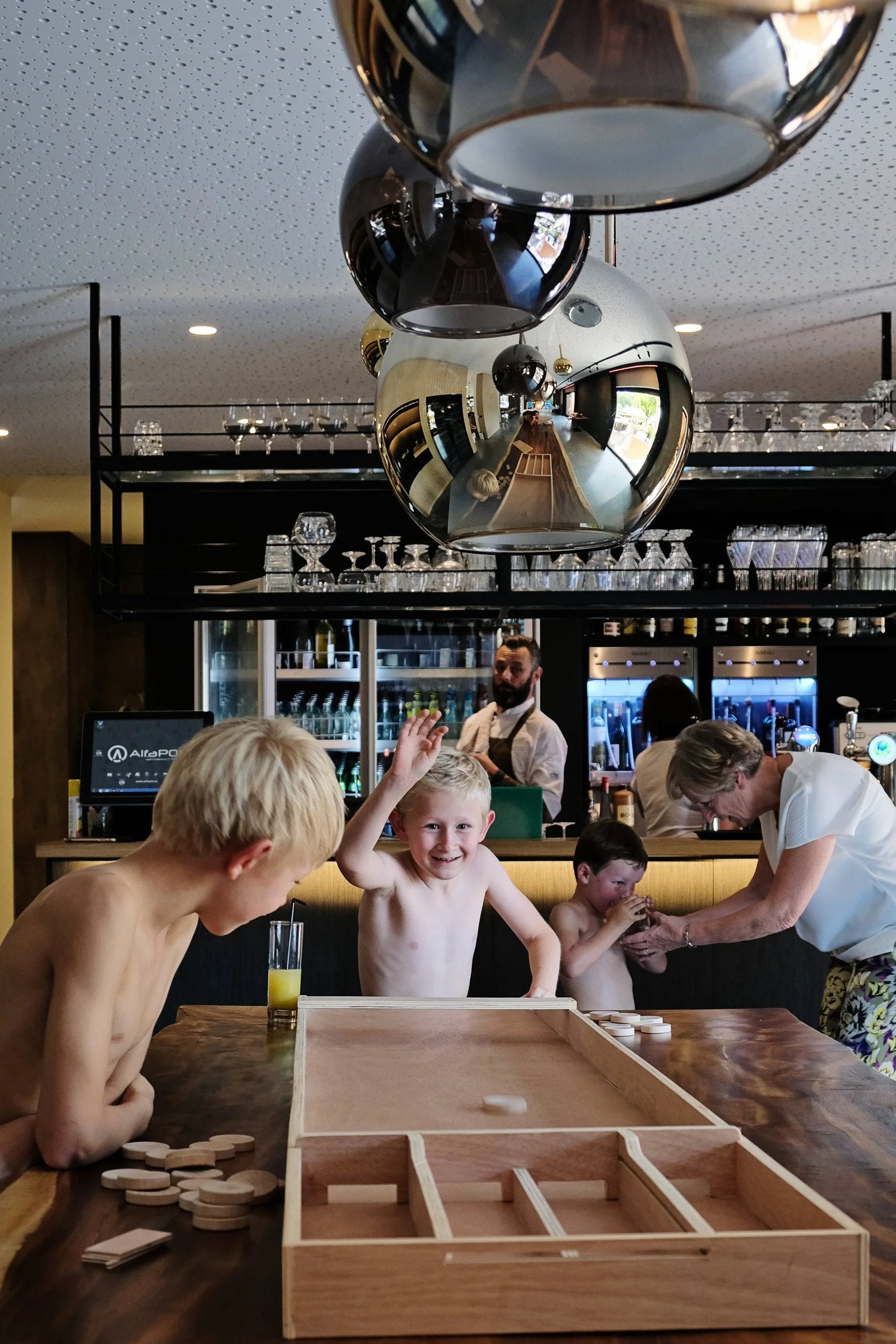 A group of children and an adult playing a wooden tabletop game in a restaurant or bar with a bar counter and hanging glasses, with reflections of the room in glossy black pendant lights above.