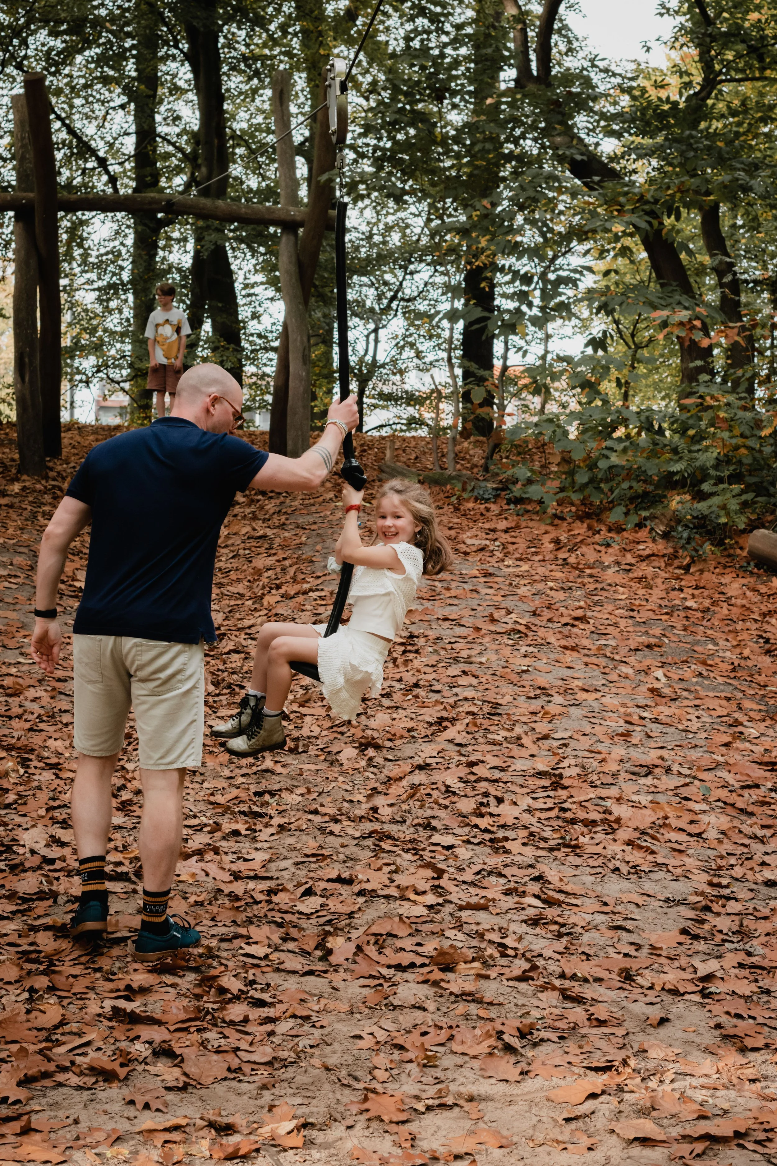 A man and a young girl on a playground in a wooded area. The girl is sitting on a black swing, holding the chain, and smiling, while the man stands nearby. In the background, a boy wearing a white shirt with a cartoon character is standing on a woode