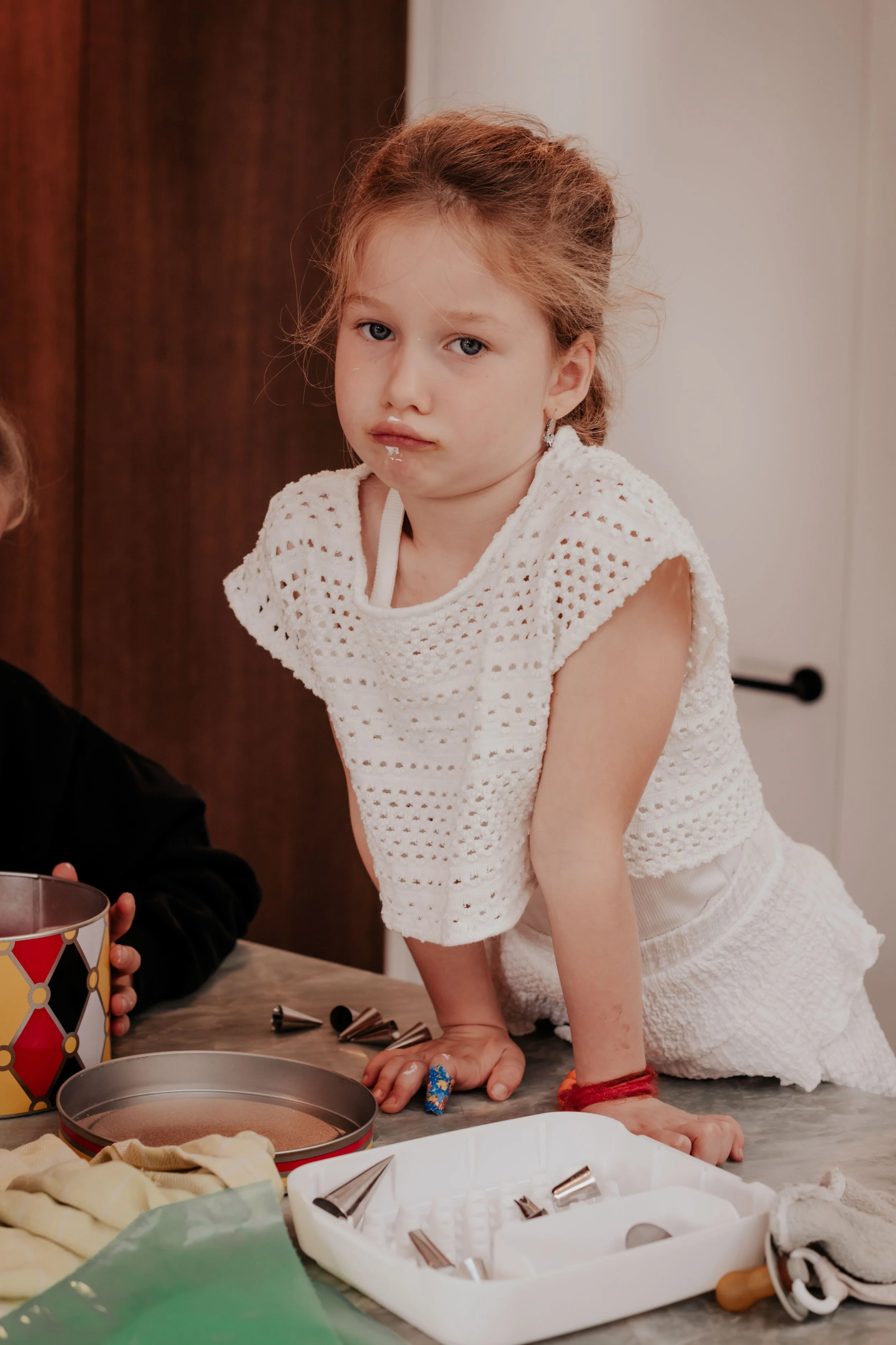 Young girl with red hair leaning on a table, surrounded by baking supplies, including cookie cutters, a baking tin, and other baking tools.