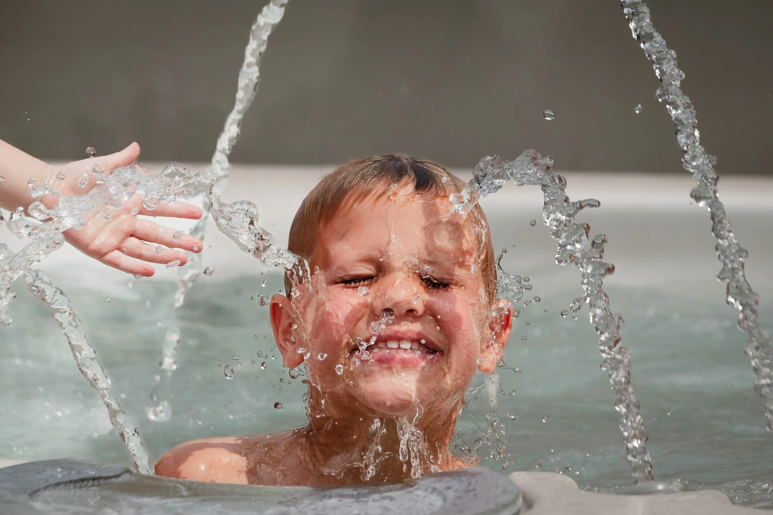 A young person with wet hair and eyes closed, smiling with water splashing around them in a swimming pool.