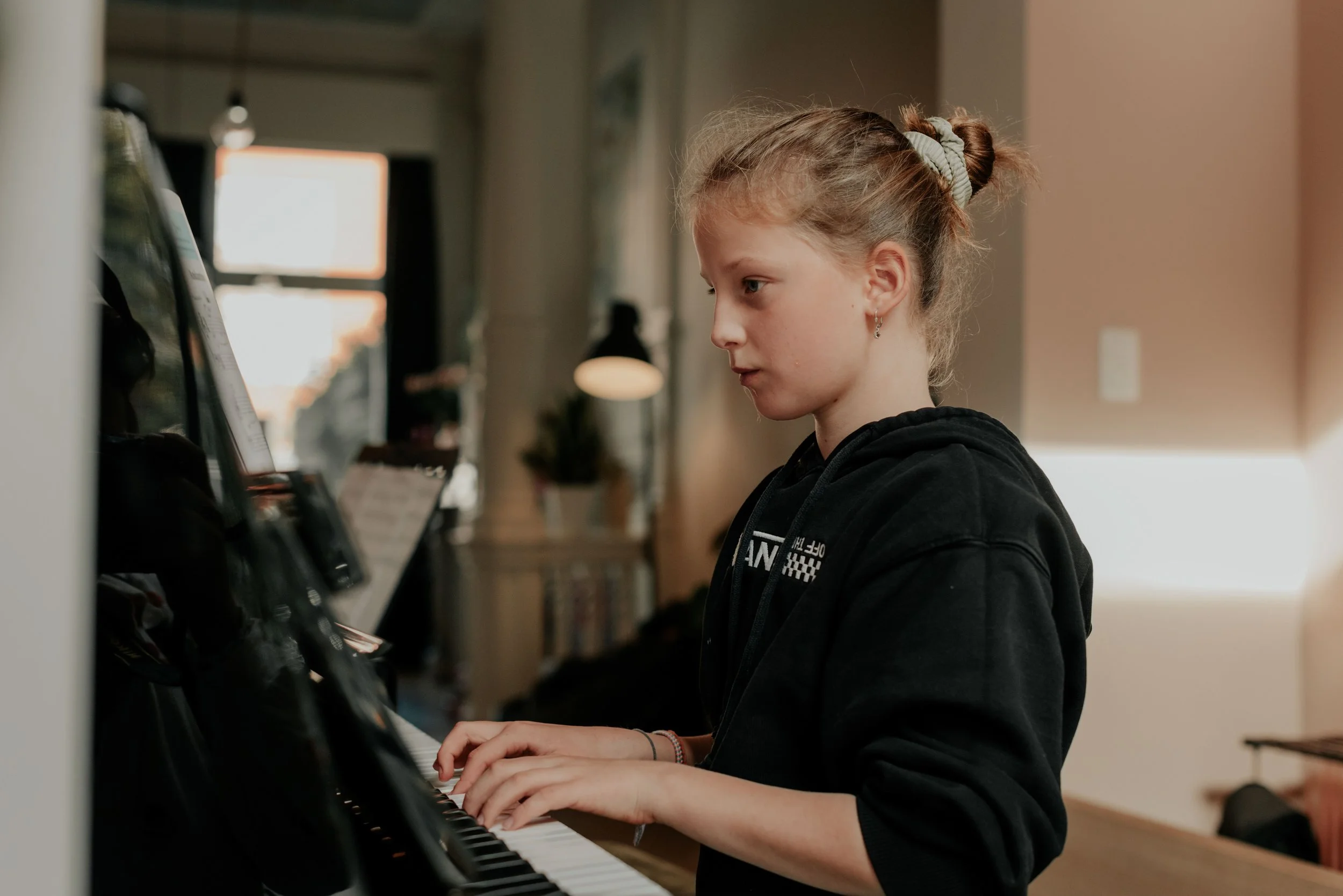 A young girl with curly hair tied in a bun and earrings playing the piano in a room with natural light from a window.