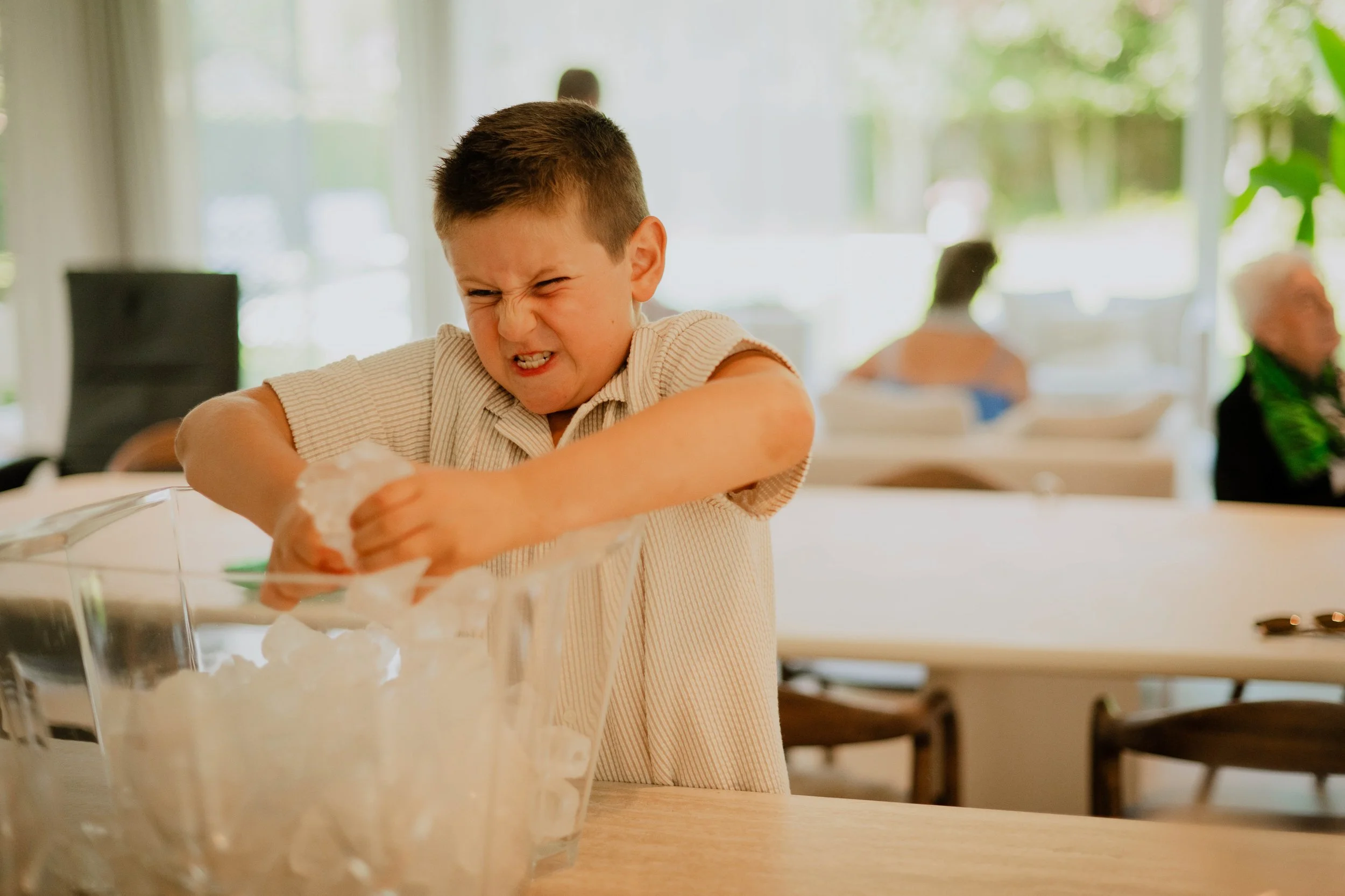 A young boy with short brown hair is angry or frustrated, sitting at a table and tearing paper. There are blurred people in the background and a glass bowl with crumpled paper in the foreground.
