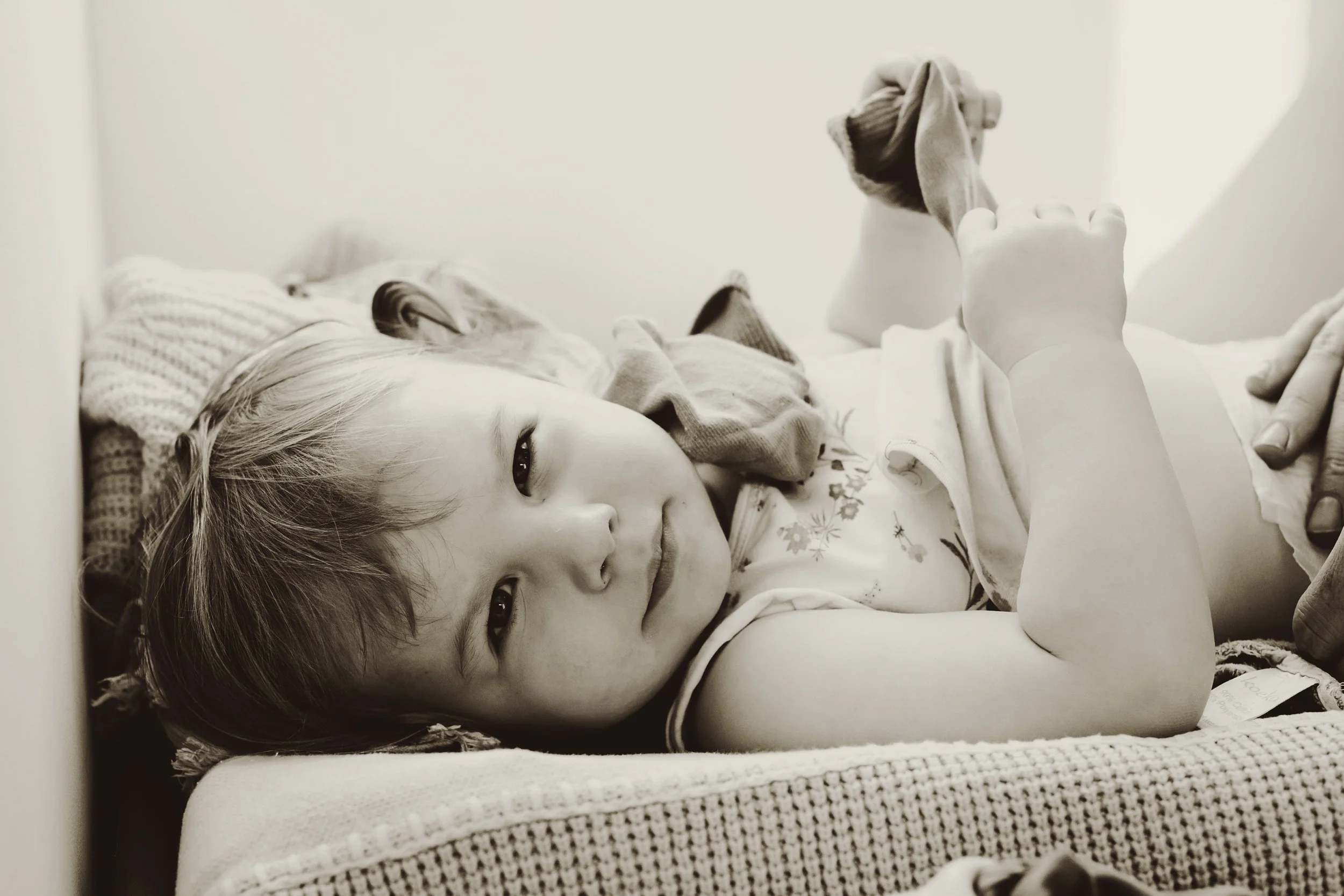 A young girl lying down inside a hospital bed, looking at the camera, with one eye open, holding a piece of cloth. She appears to be in a medical setting.