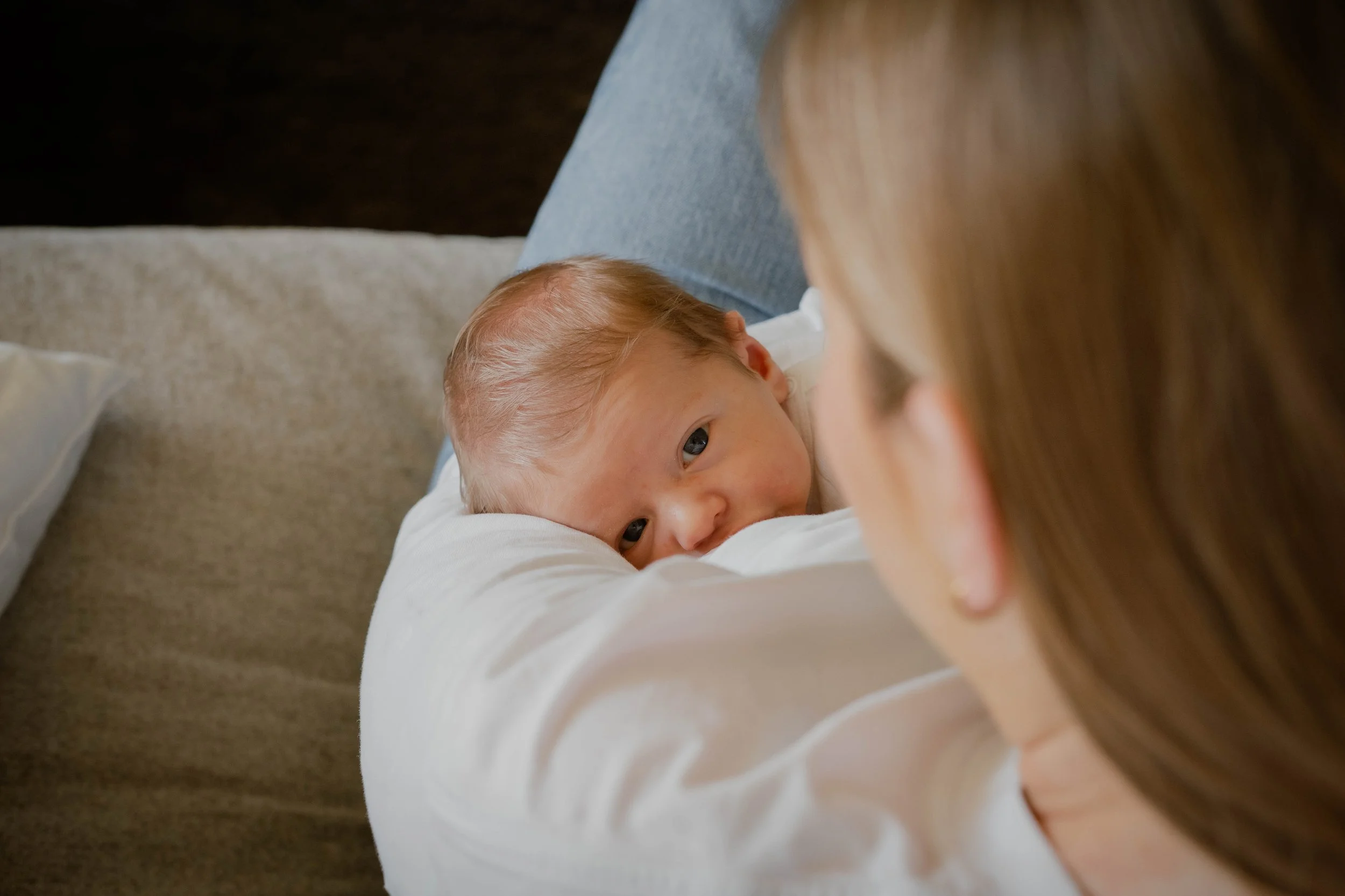 A woman with long blonde hair holding a newborn baby with light skin and blue eyes, lying on a beige sofa.