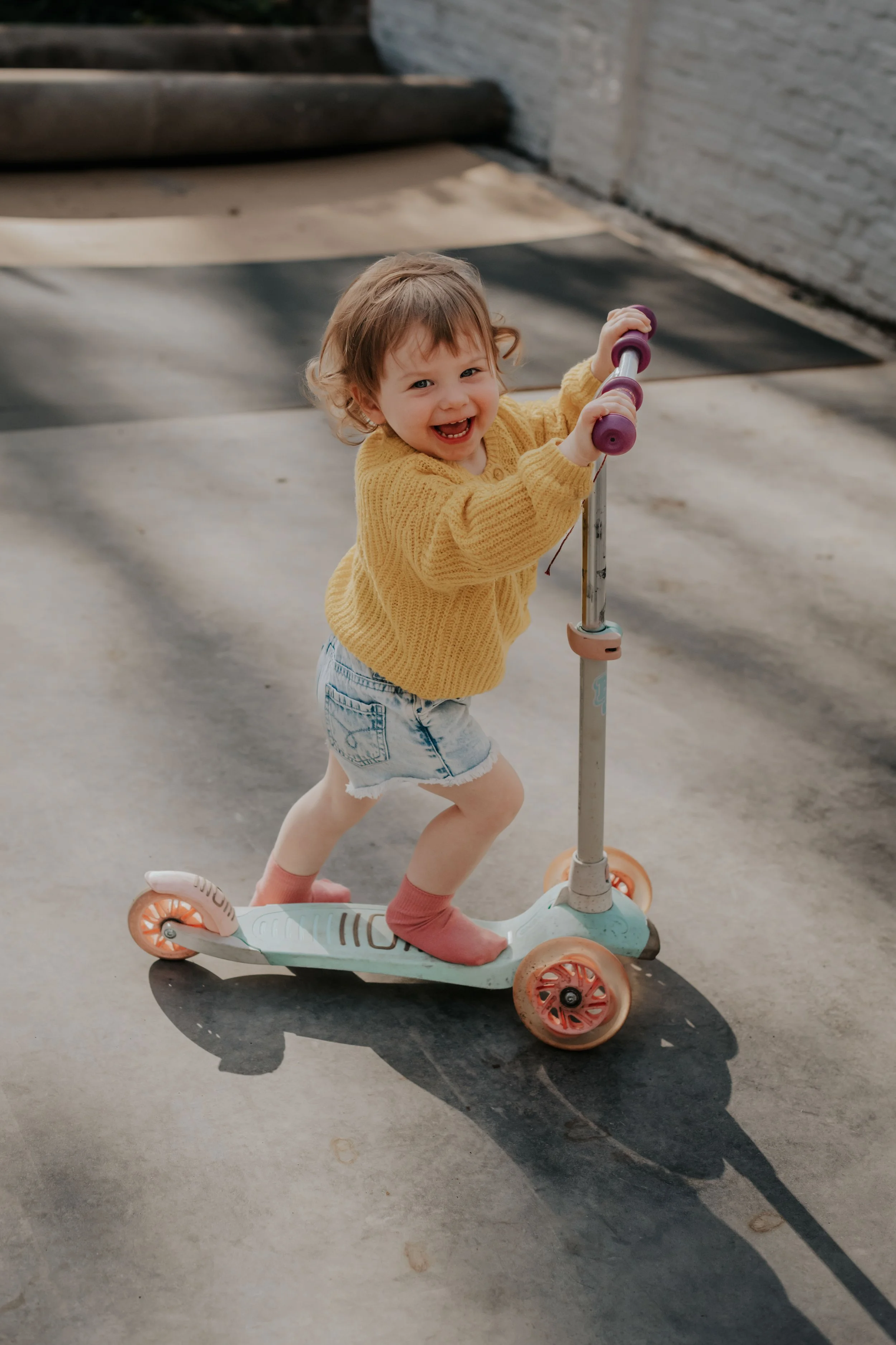 A young girl with short, curly hair and a big smile riding a light blue and coral scooter on a concrete surface, wearing a yellow sweater, denim shorts, pink socks, and white shoes.
