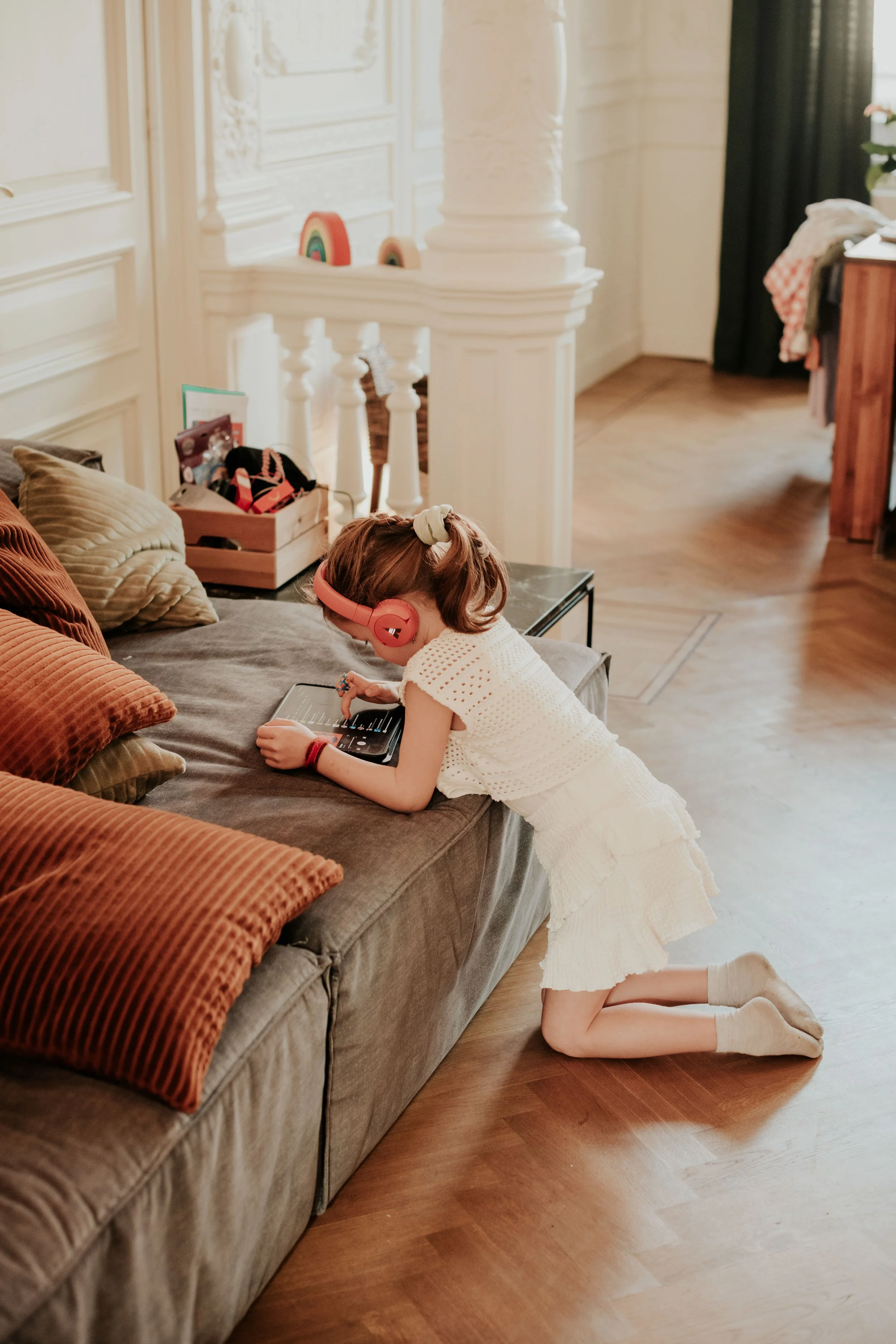 A young girl with a cream dress and socks, wearing pink headphones, is kneeling on the floor and playing with a tablet on a gray couch in a cozy living room.