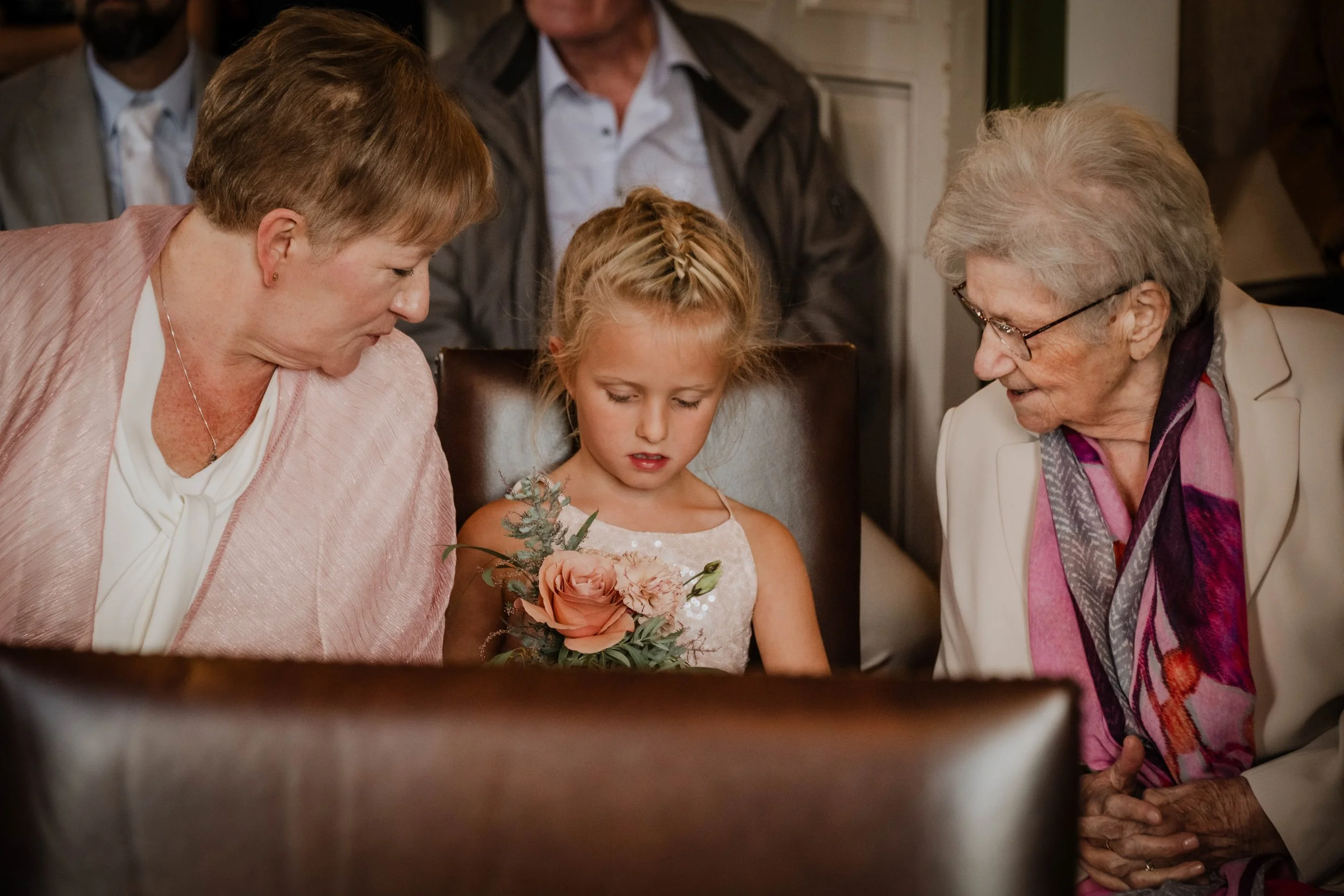 A young girl holding a bouquet of flowers is standing between two elderly women, all three leaning in closely and looking at something in front of them, possibly a display or a gift, inside a room that appears to be a home or venue.
