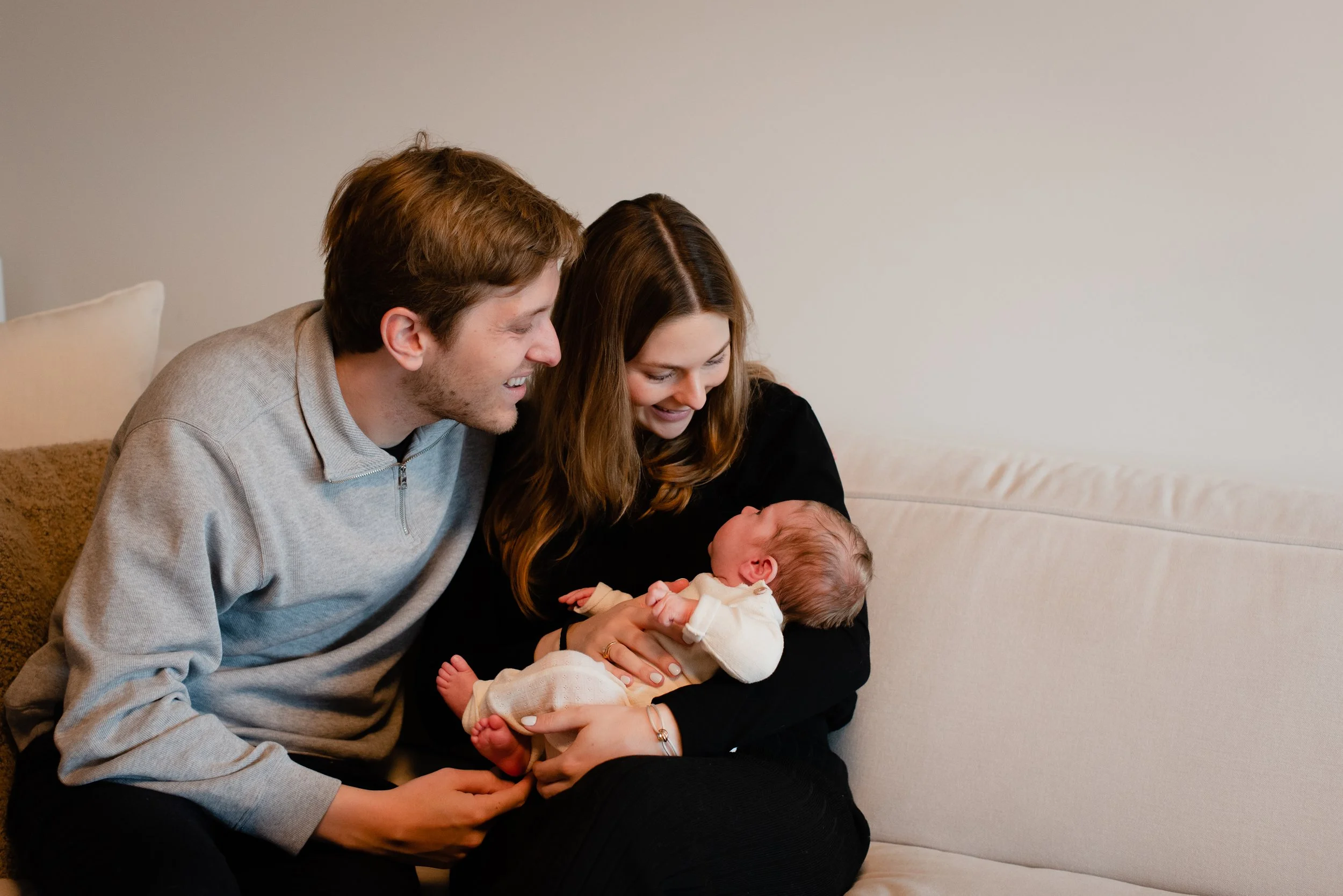 A happy couple holds their newborn baby on a beige sofa in a living room.
