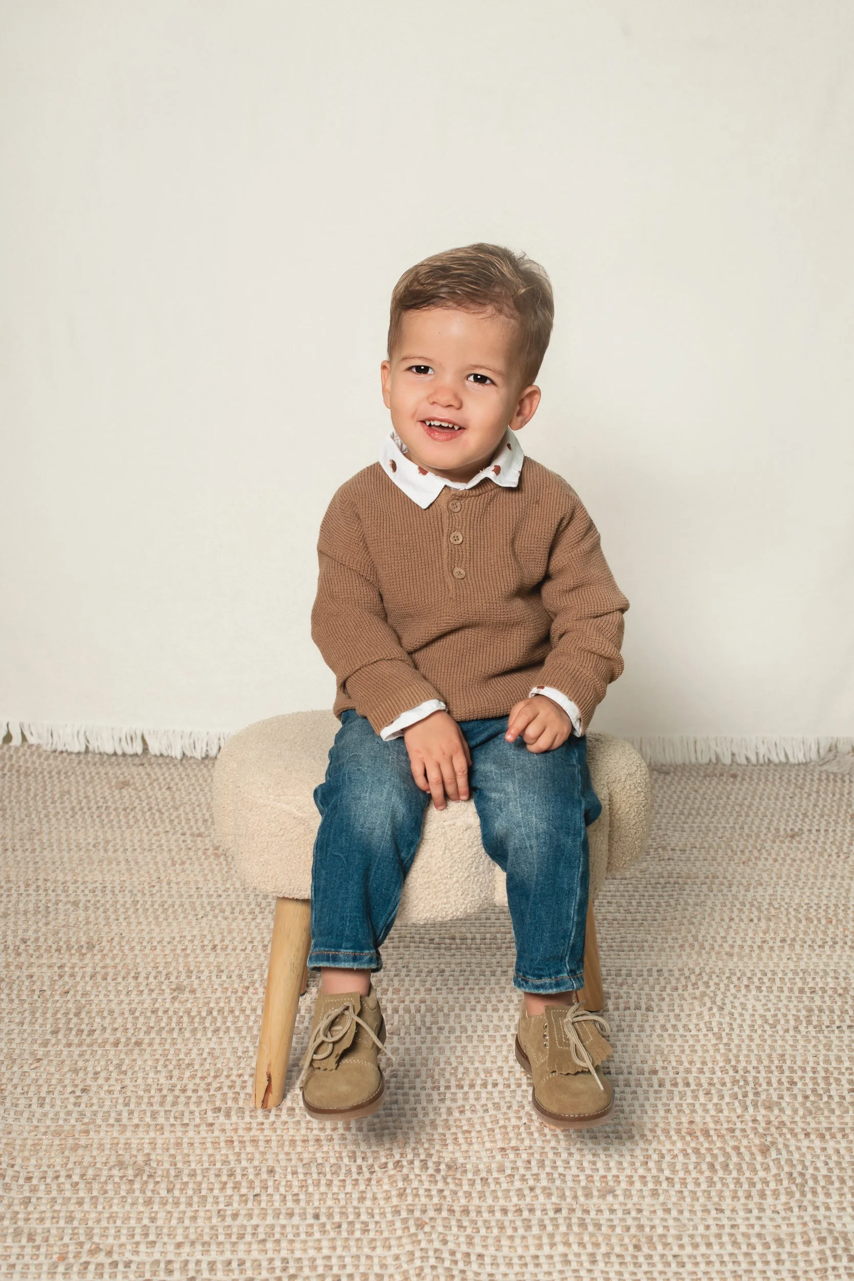 A young child sitting on a beige stool, wearing a brown sweater, white button-up shirt, blue jeans, and tan boots, smiling with a slight tilt of the head, against a plain light-colored background.