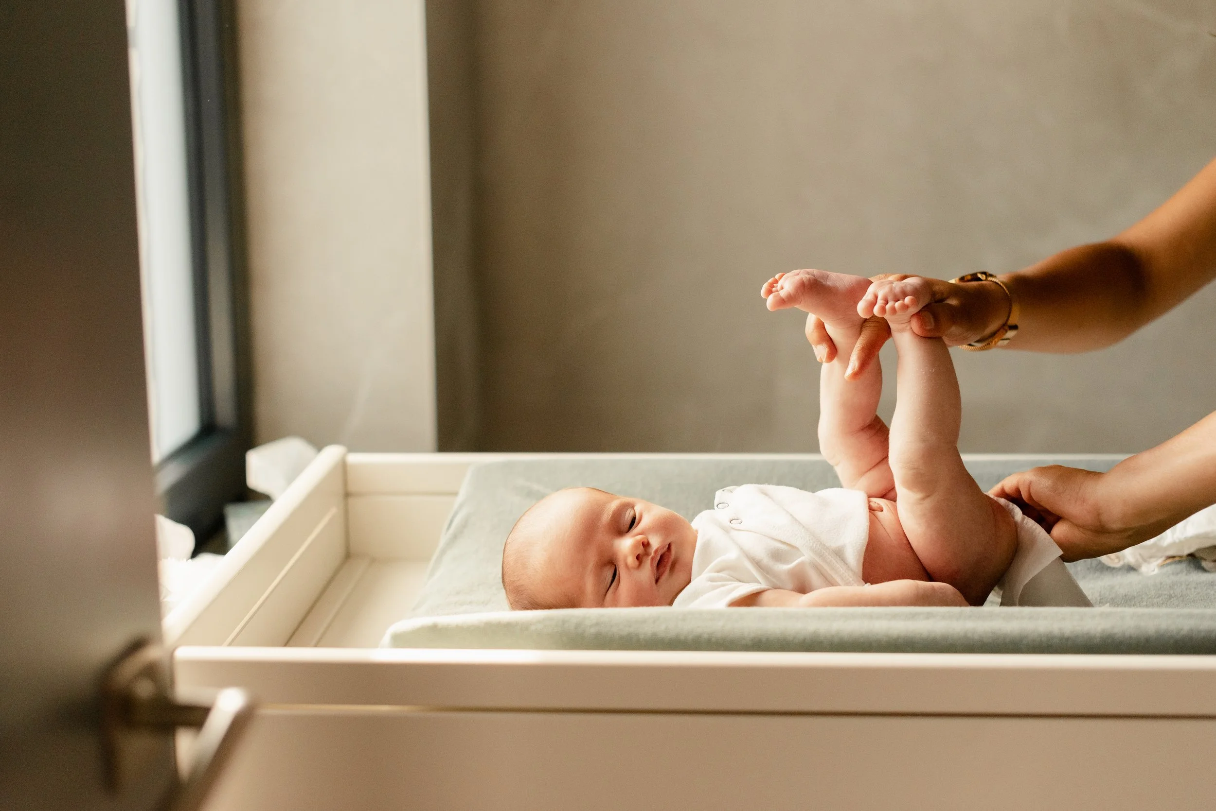 A baby lying in a hospital bassinet, holding an adult's finger, with an adult touching the baby's hand, in a hospital room.