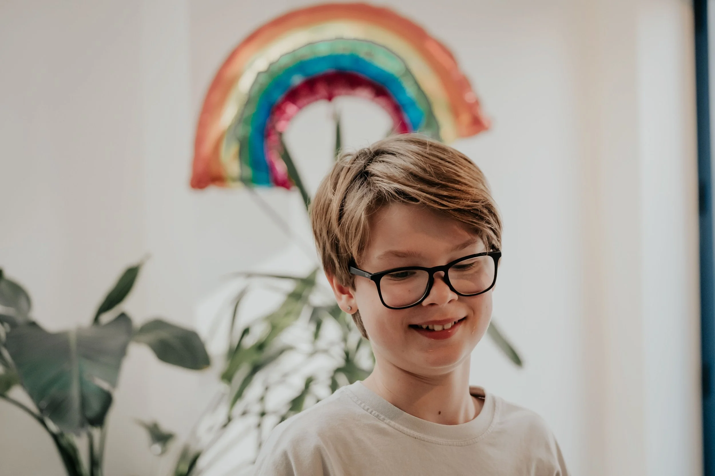 A smiling person with short hair wearing glasses and a white shirt, with a colorful rainbow decoration on the wall in the background, and greenery in the foreground.